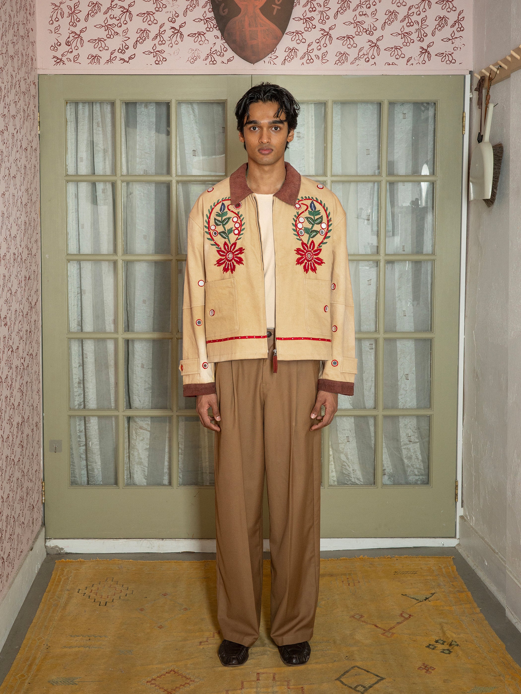 Indoors, a person wears tan trousers and the Found Paisley Motif Hand Embroidered Work Jacket featuring red floral details, paired with a light shirt, standing on a yellow rug by a glass-paneled door with lace curtains.