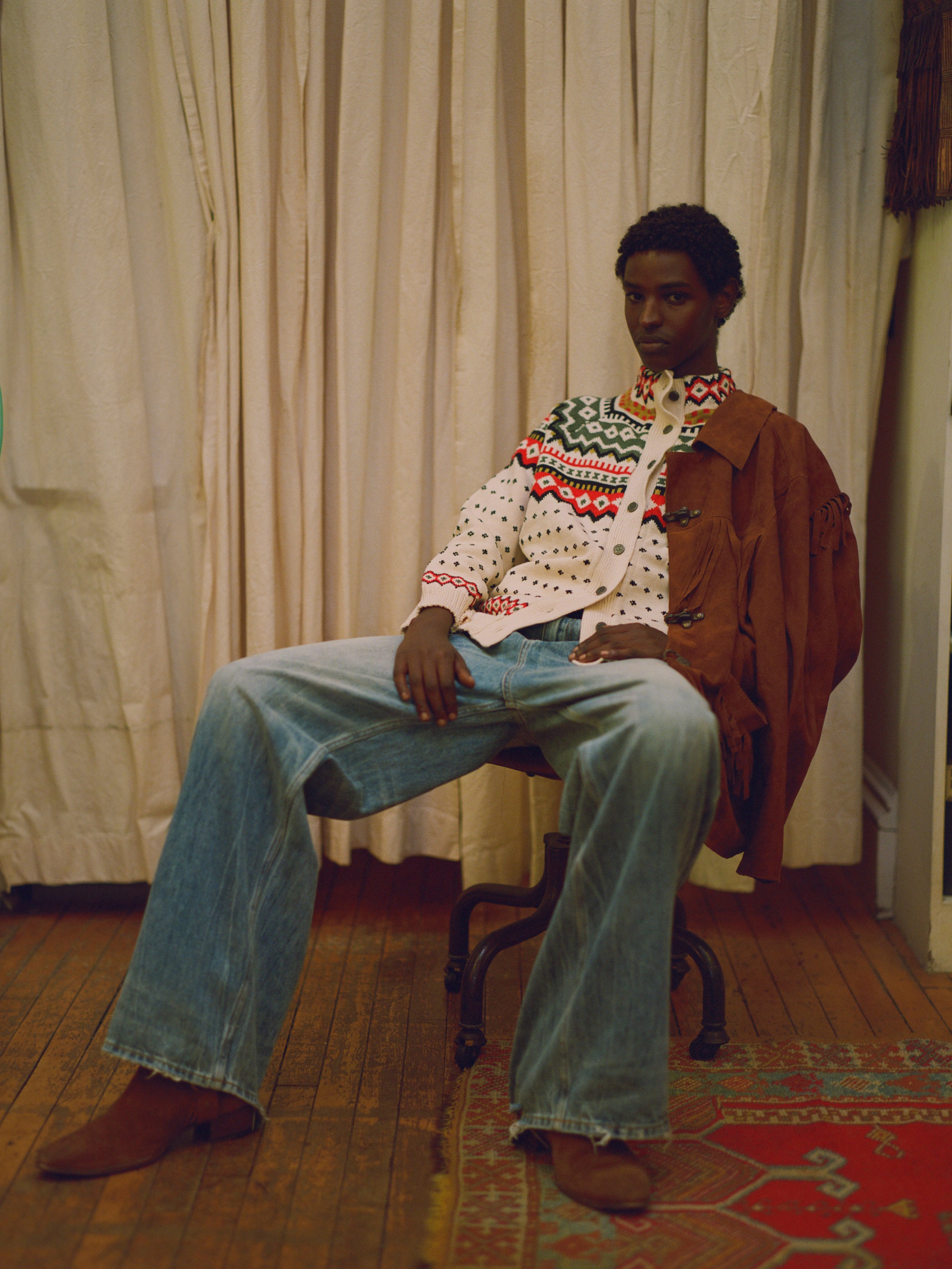 A man, wearing a cozy Harwan Isles Sweater from the Scottish Fair Isle region, sits comfortably on a chair adorned with intricate patterns.