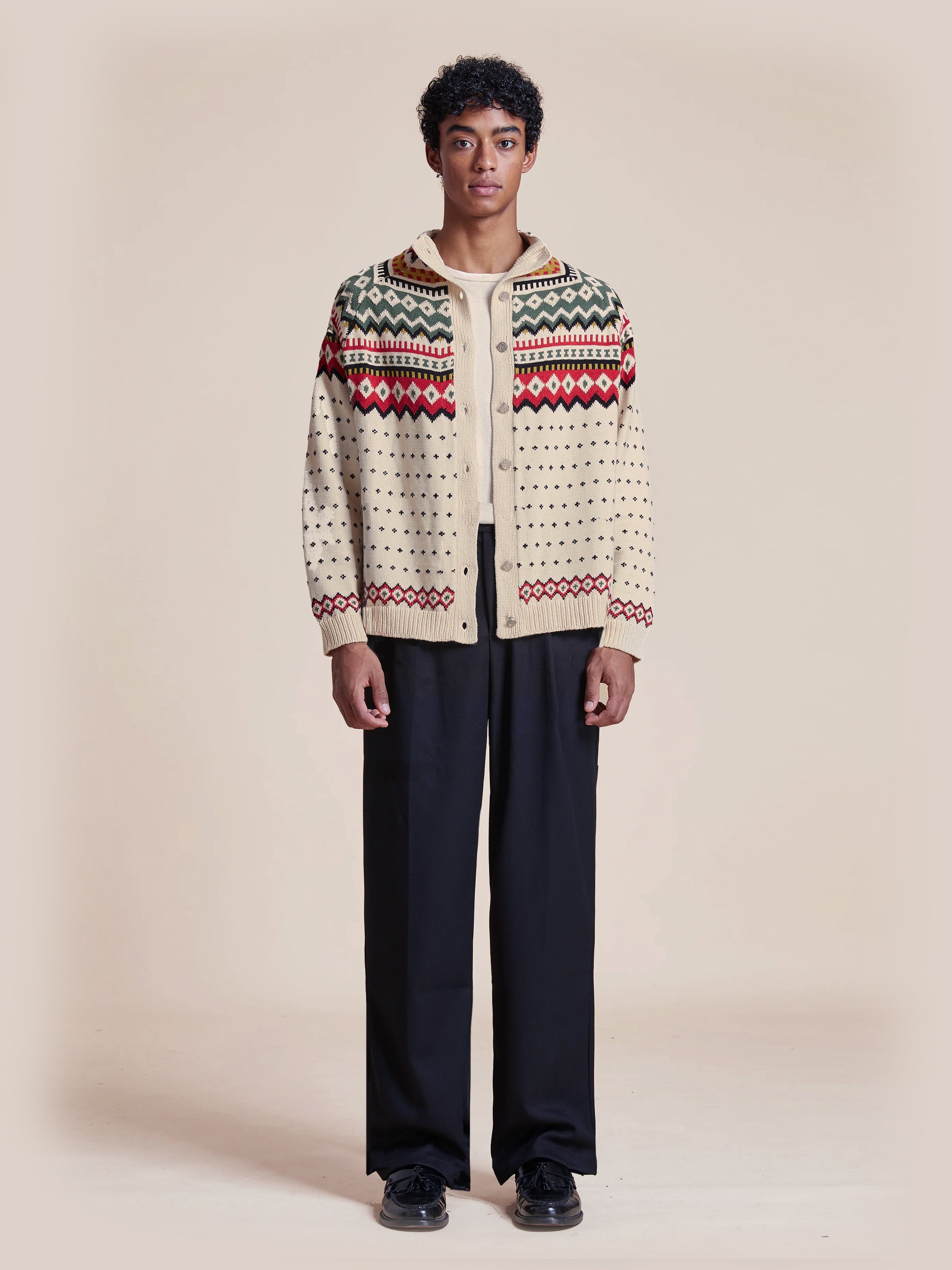 A person wears the Harwan Isles Sweater by Found, featuring classic red and green Fair Isle patterns, paired with black pants and shoes, against a plain background.