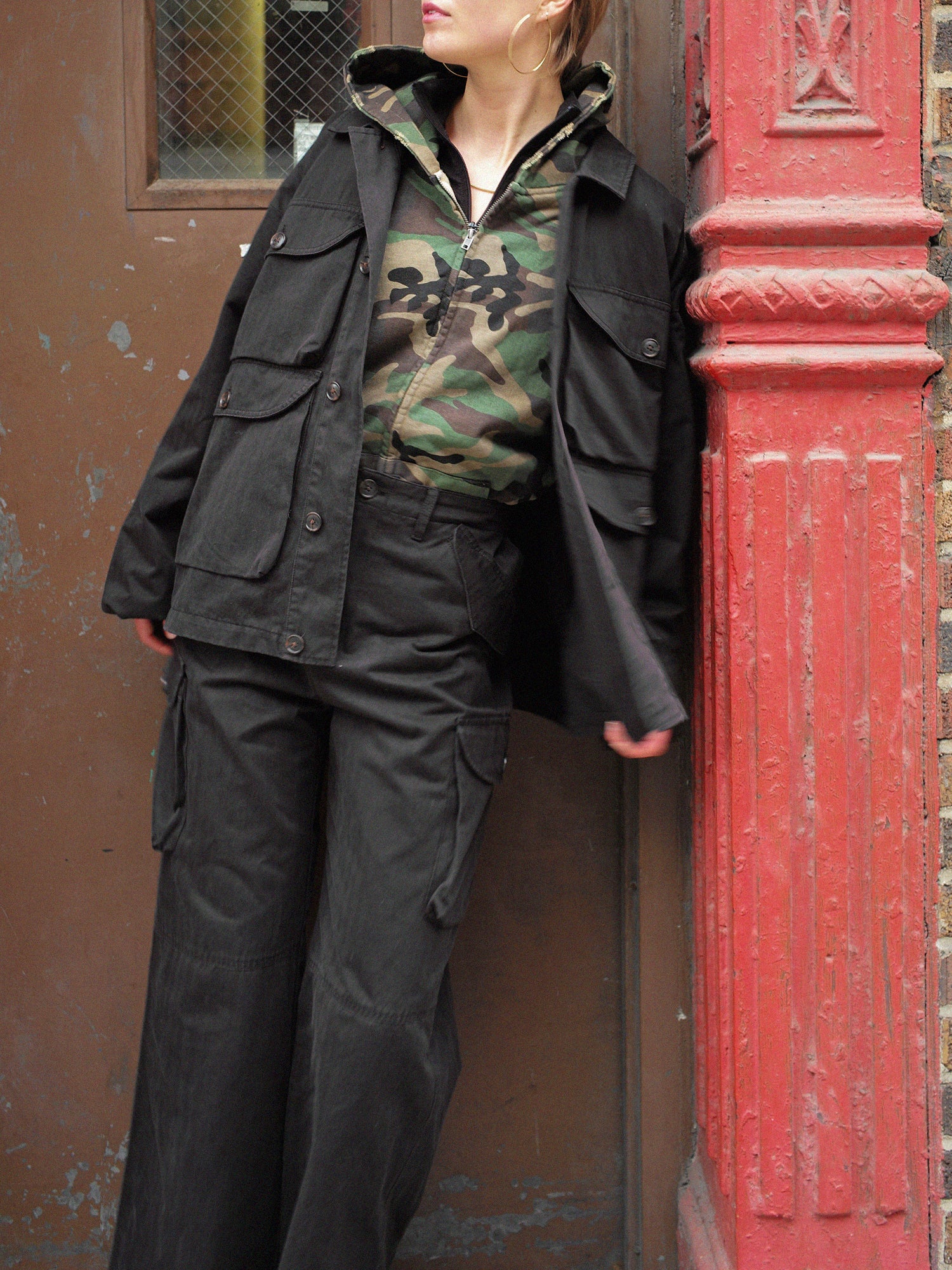 A person in a camouflage hoodie and a FOUND Utility Herringbone Cargo Jacket leans against a red textured pillar beside a brown door.