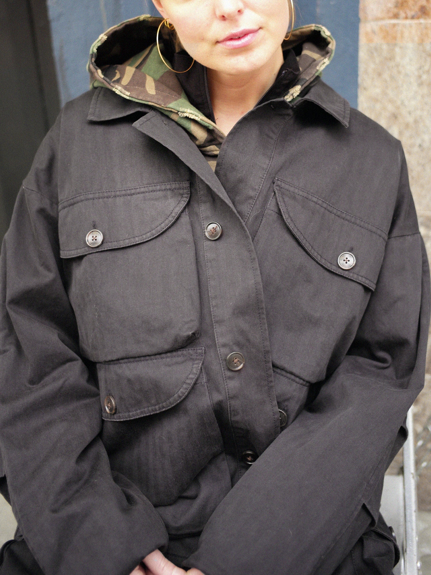 Someone wearing a dark Utility Herringbone Cargo Jacket by FOUND over a camouflage hoodie, showcasing a stylish unisex relaxed fit.