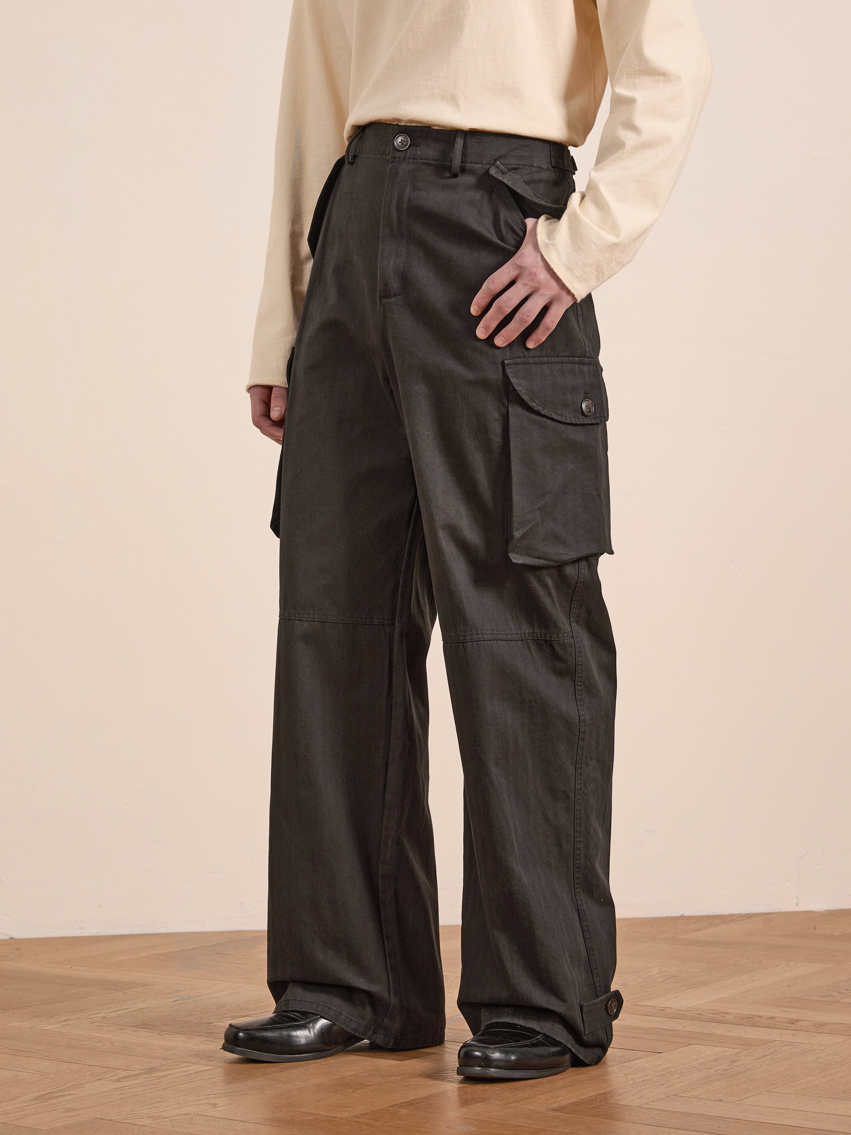 A person wearing a beige long-sleeve shirt and dark Utility Herringbone Cargo Pants by FOUND stands on a wooden floor, exuding a vintage look.