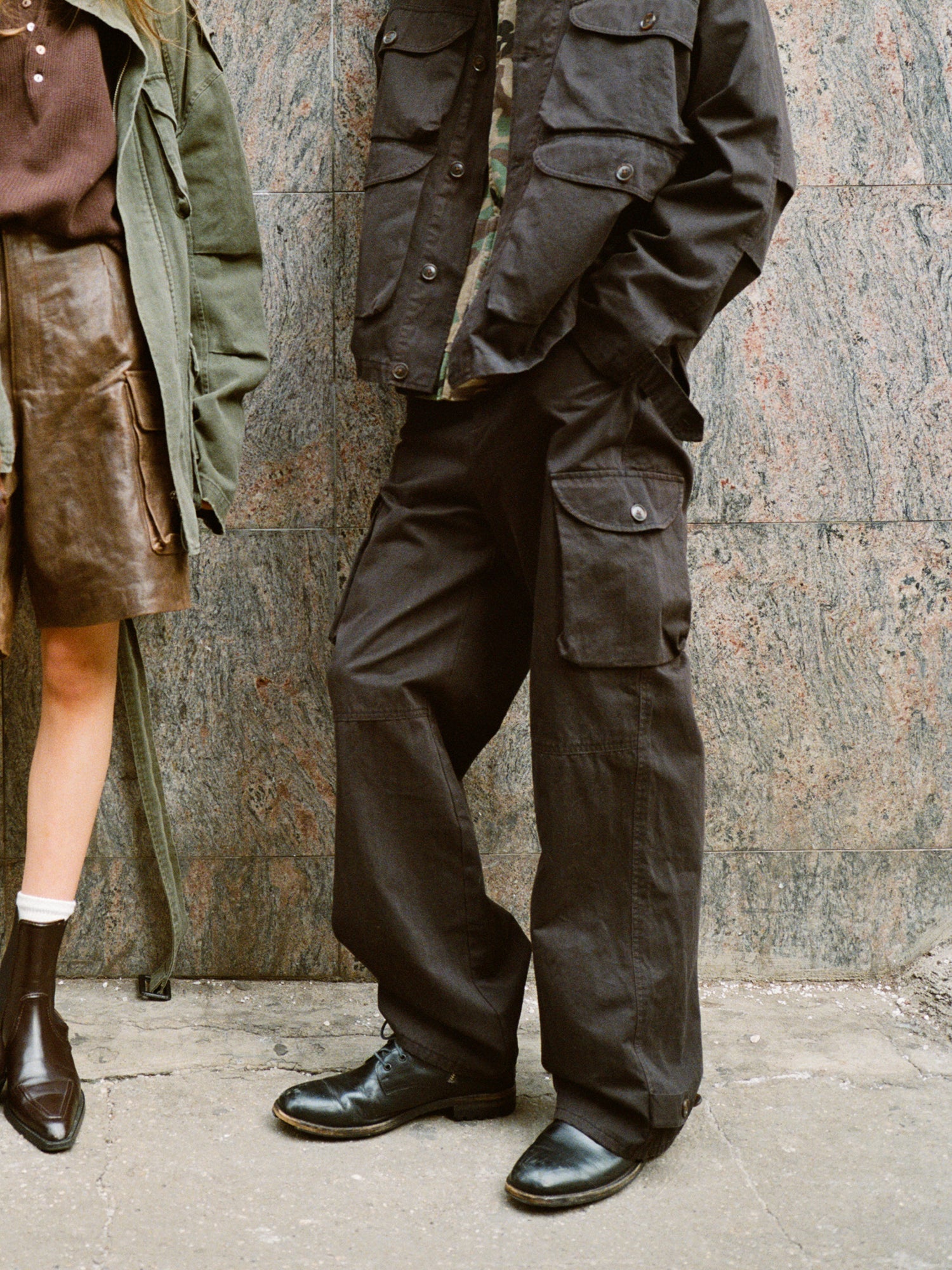 Two people stand against a textured wall. One wears a skirt and jacket, while the other sports FOUND's Utility Herringbone Cargo Pants with a relaxed fit, paired perfectly with their jacket.