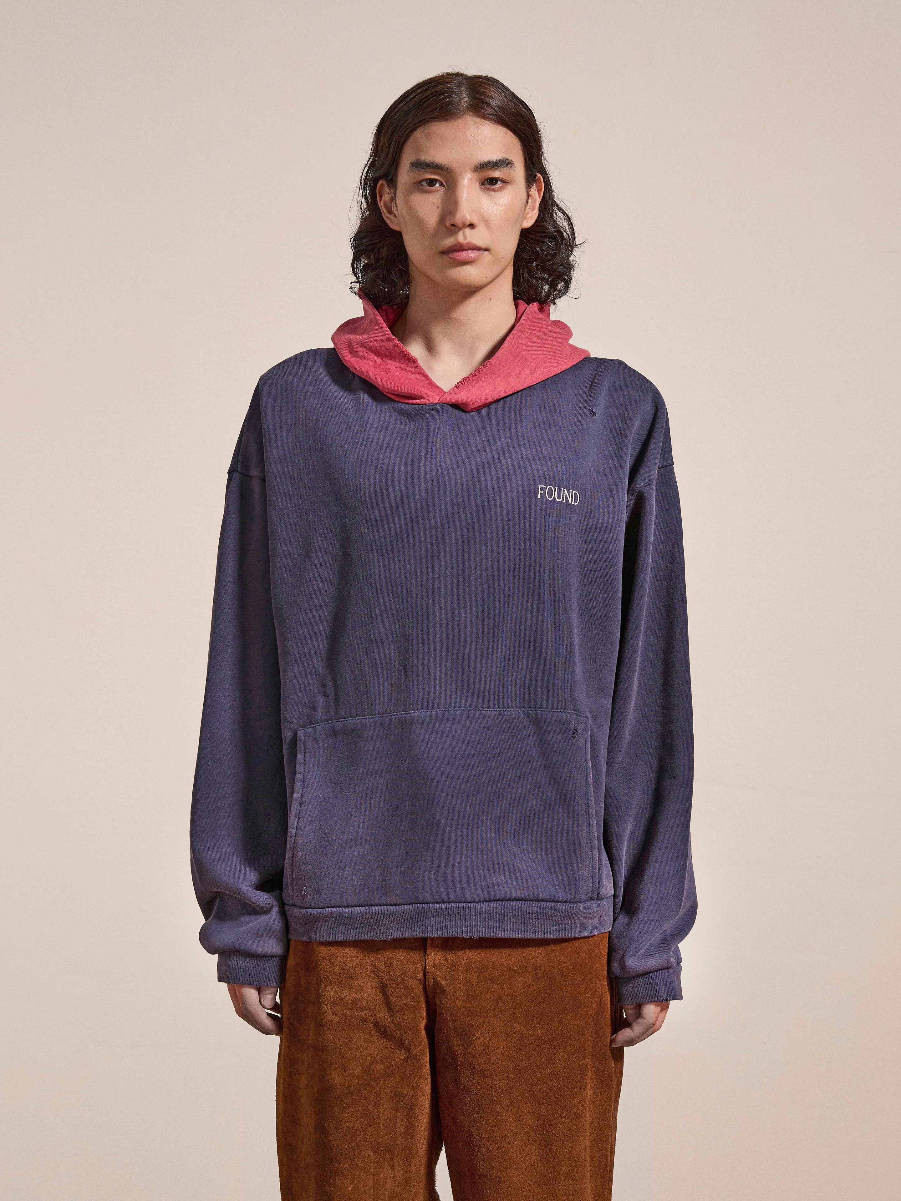 A person with long hair stands against a plain background, wearing the FOUND Distressed Logo Hoodie. The blue hoodie, made from French terry cotton, features a red hood and the word "FOUND" displayed in a distressed logo style on it. They are paired with brown trousers.