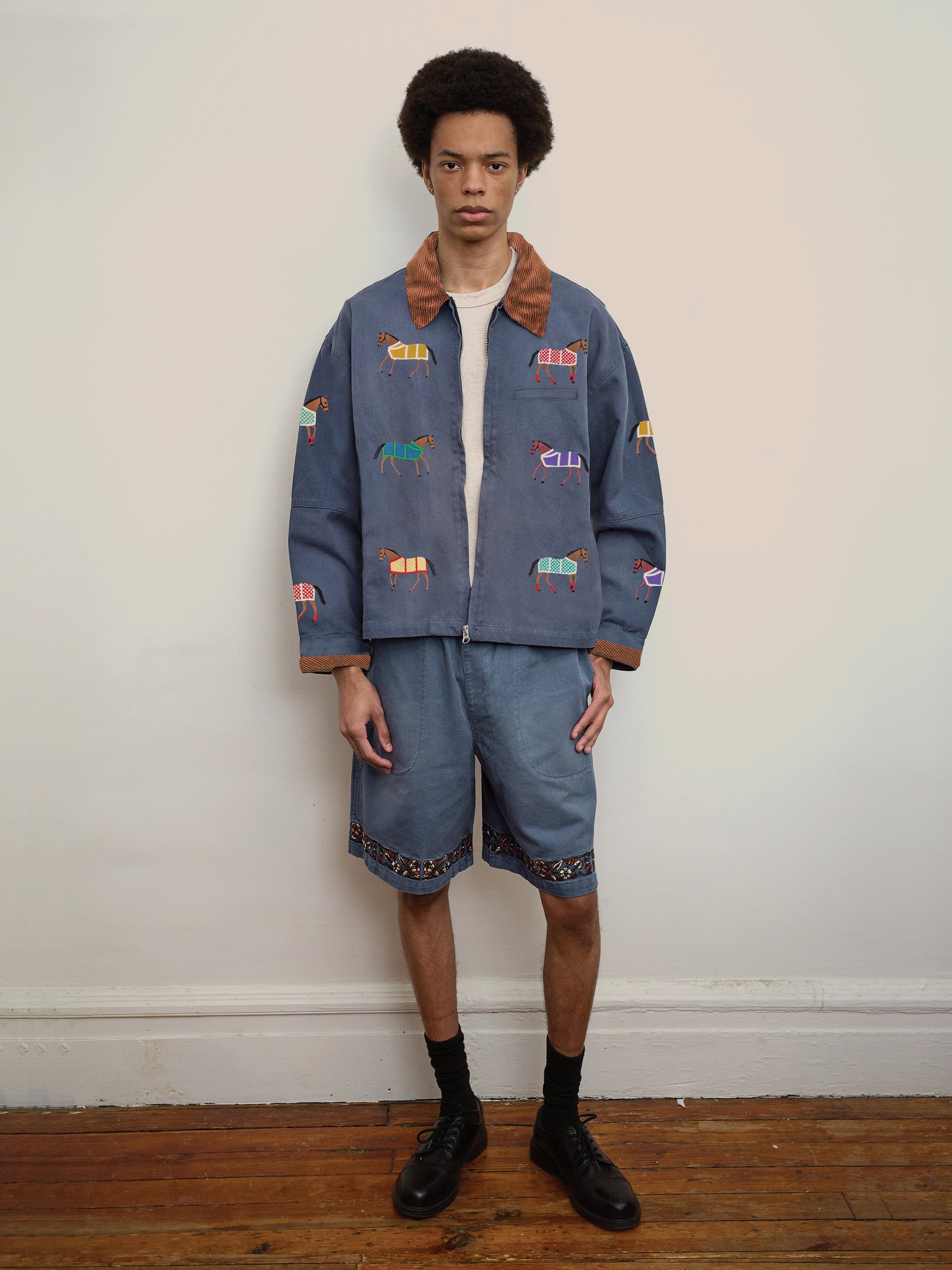 A person stands indoors against a plain wall wearing the Found Lariat Horse Embroidered Work Jacket, matching blue shorts, a white t-shirt, black socks, and black shoes.