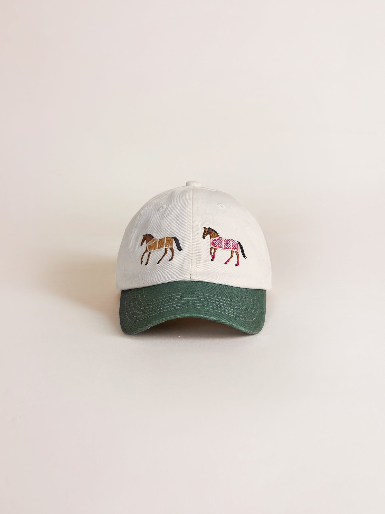 The Horse Equine Cap from Found is a white baseball cap with a green brim, featuring two embroidered horses on the front: one brown and the other pink with a patterned saddle that evokes retro 70's color-blocking.