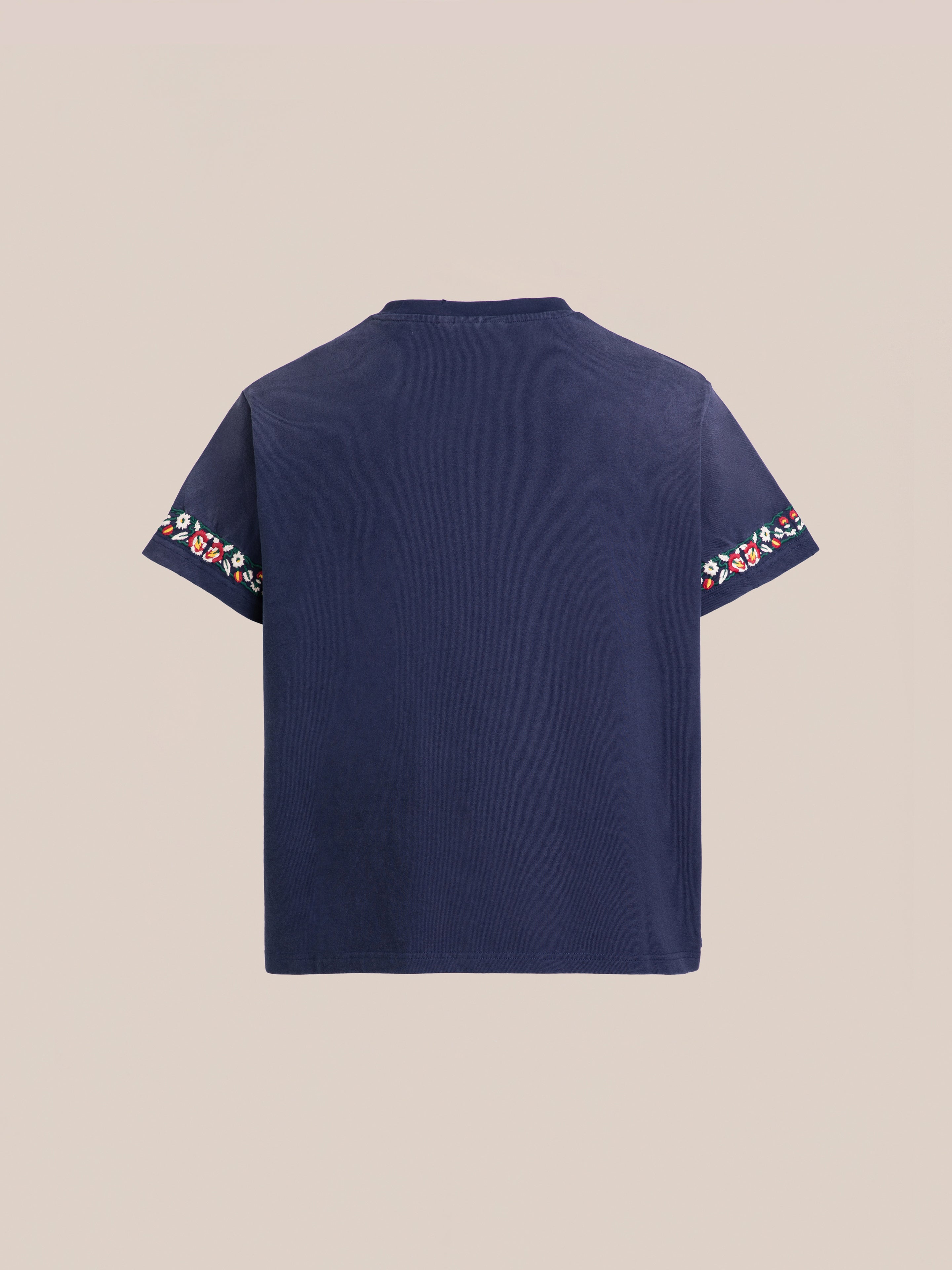 The Horse Equine Tee by FOUND is a navy blue T-shirt with short sleeves, adorned with colorful embroidered floral patterns on the ends of each sleeve. The background is beige.