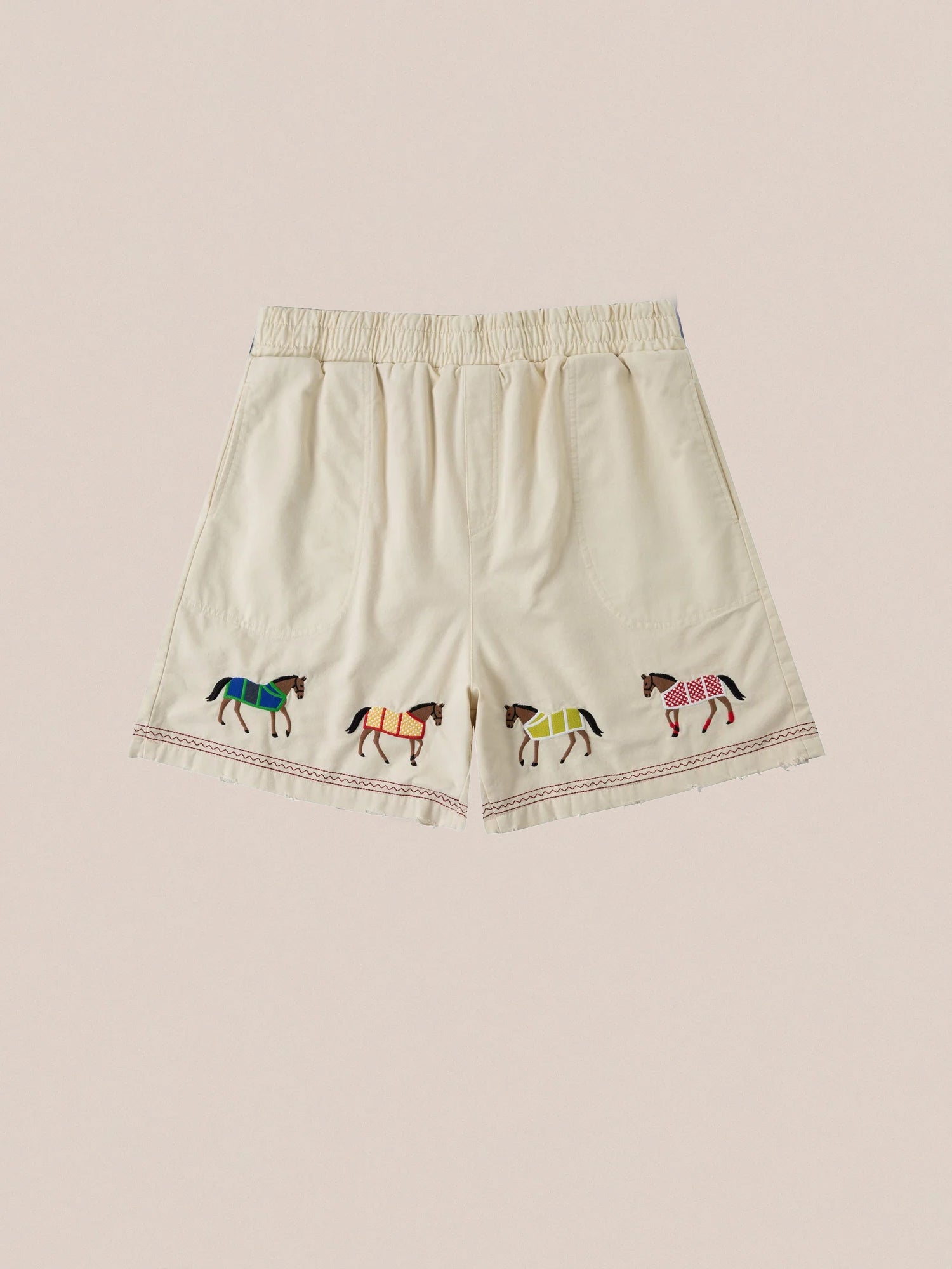 The Embroidered Horse Shorts by Profound are beige, made from cotton twill, and feature an elastic waistband and embroidered horse motifs in multicolored blankets along the hem.