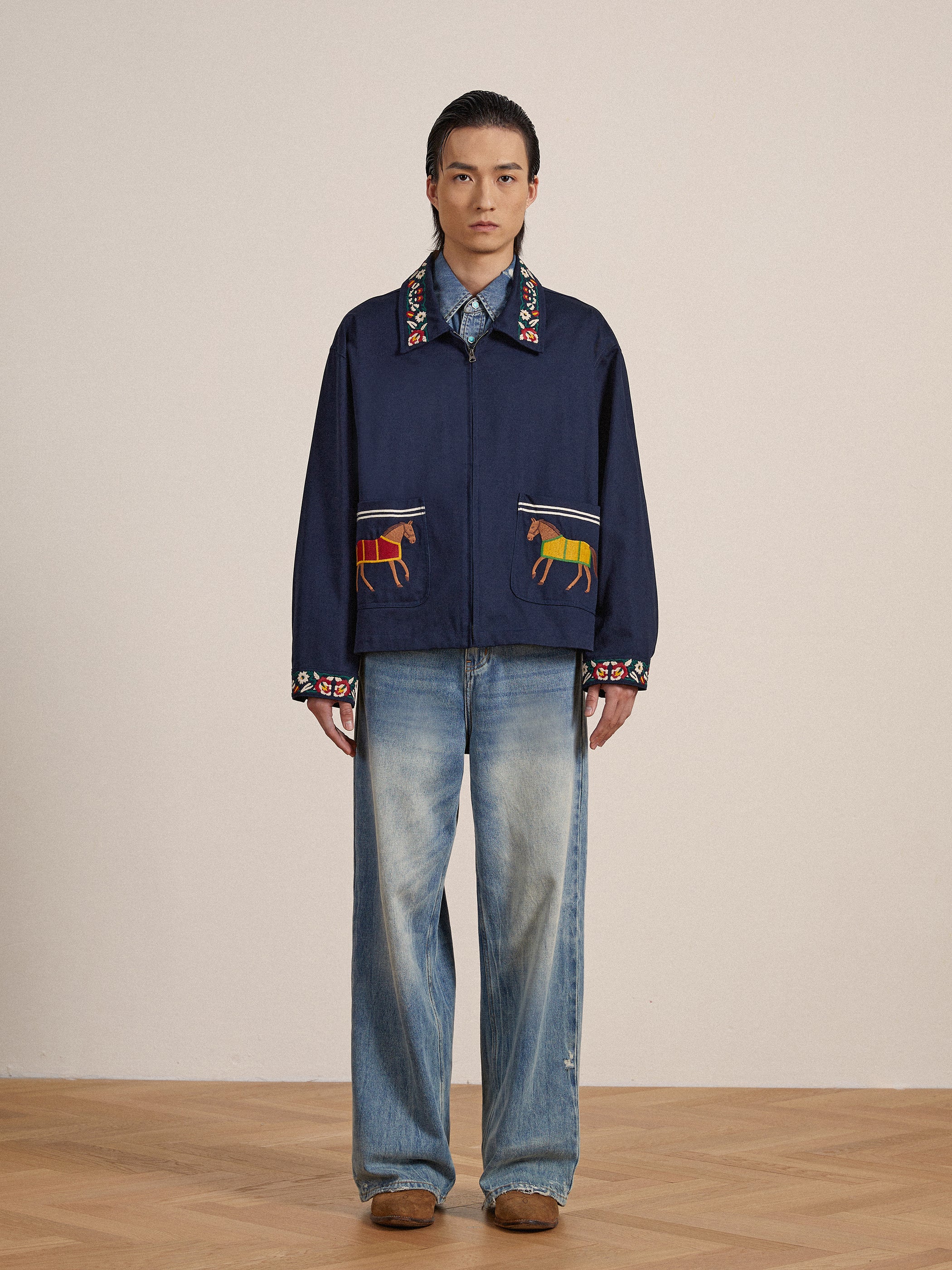 A jockey stands wearing a navy Found Horse Equine Work Jacket with colorful embroidered detail and faded jeans, against a plain beige background.