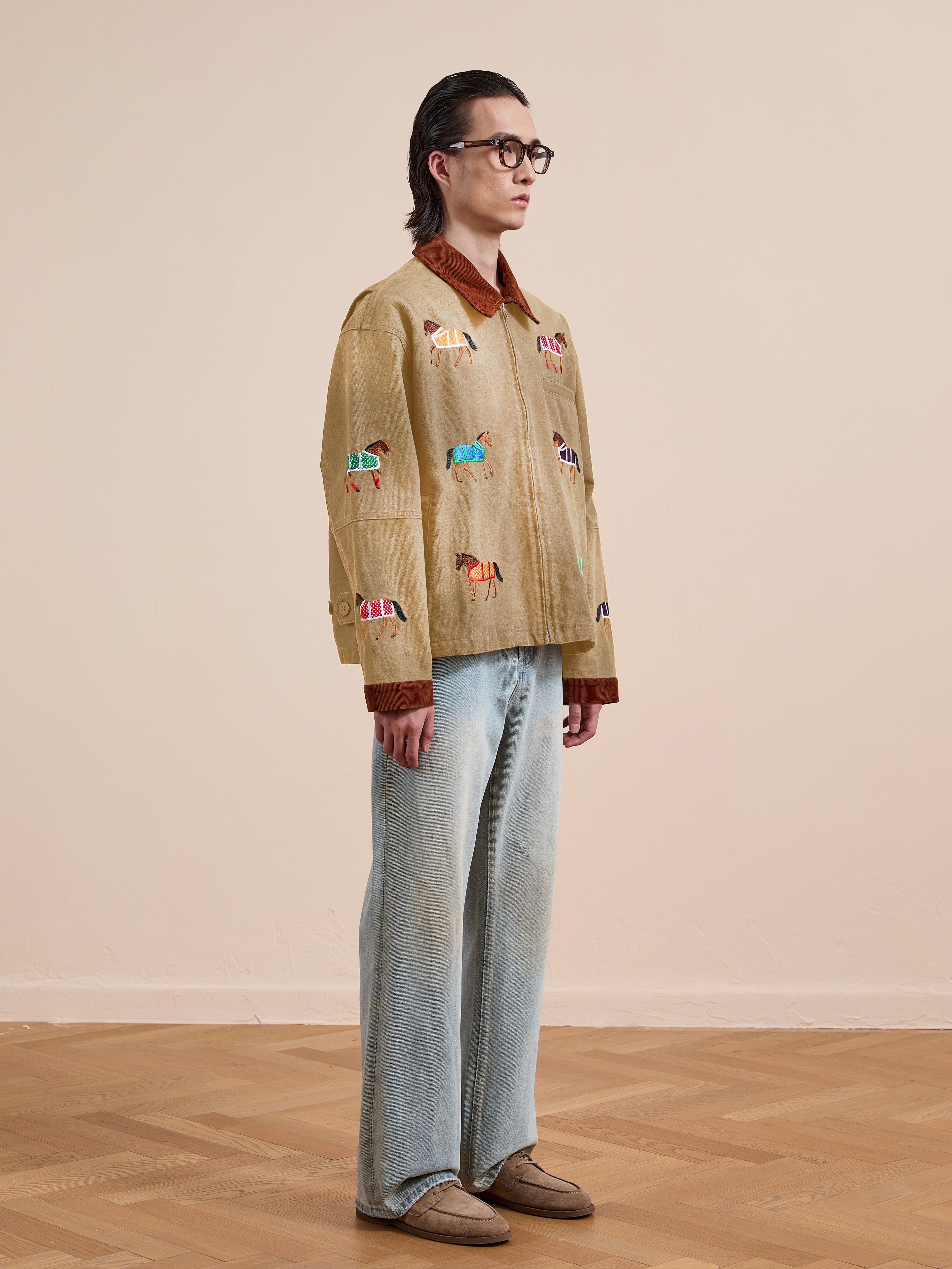 A person stands indoors wearing glasses, a brown shirt, light blue jeans, beige shoes, and the Found Lariat Horse Embroidered Work Jacket, which is tan with colorful horse designs, facing left.