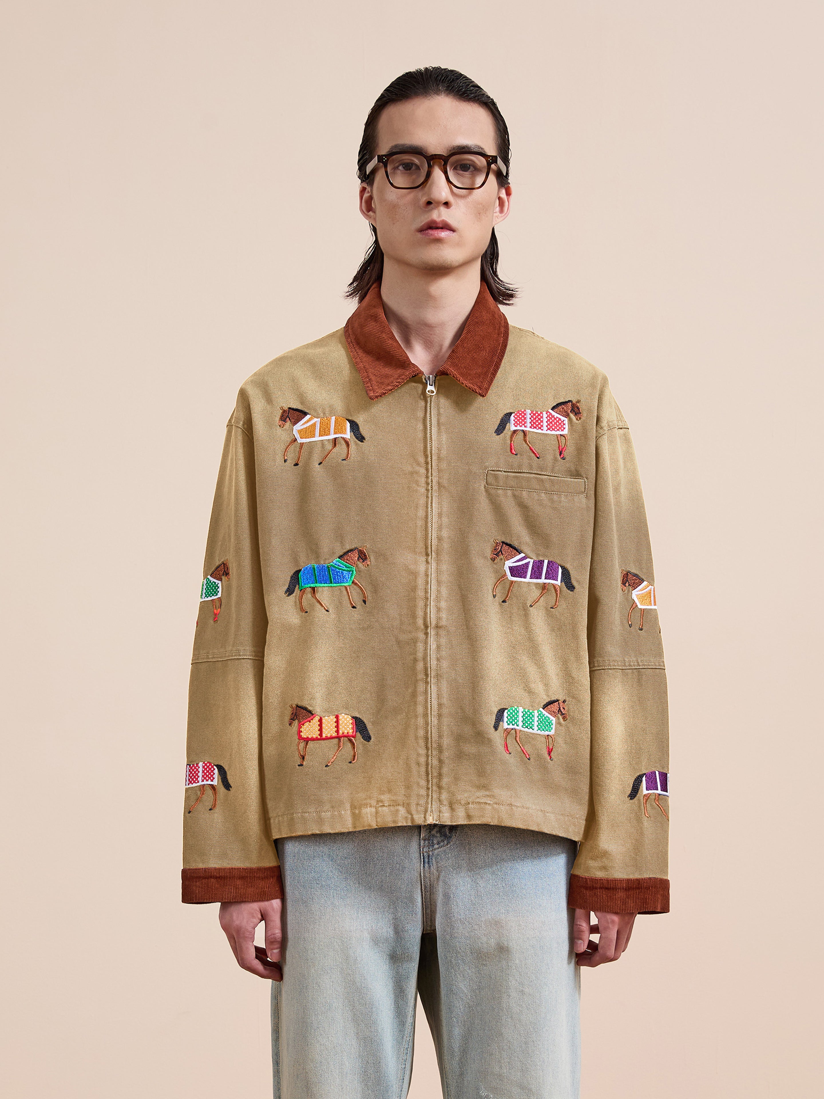 A person wearing glasses and the Found Lariat Horse Embroidered Work Jacket, paired with light blue jeans, stands against a plain beige background.