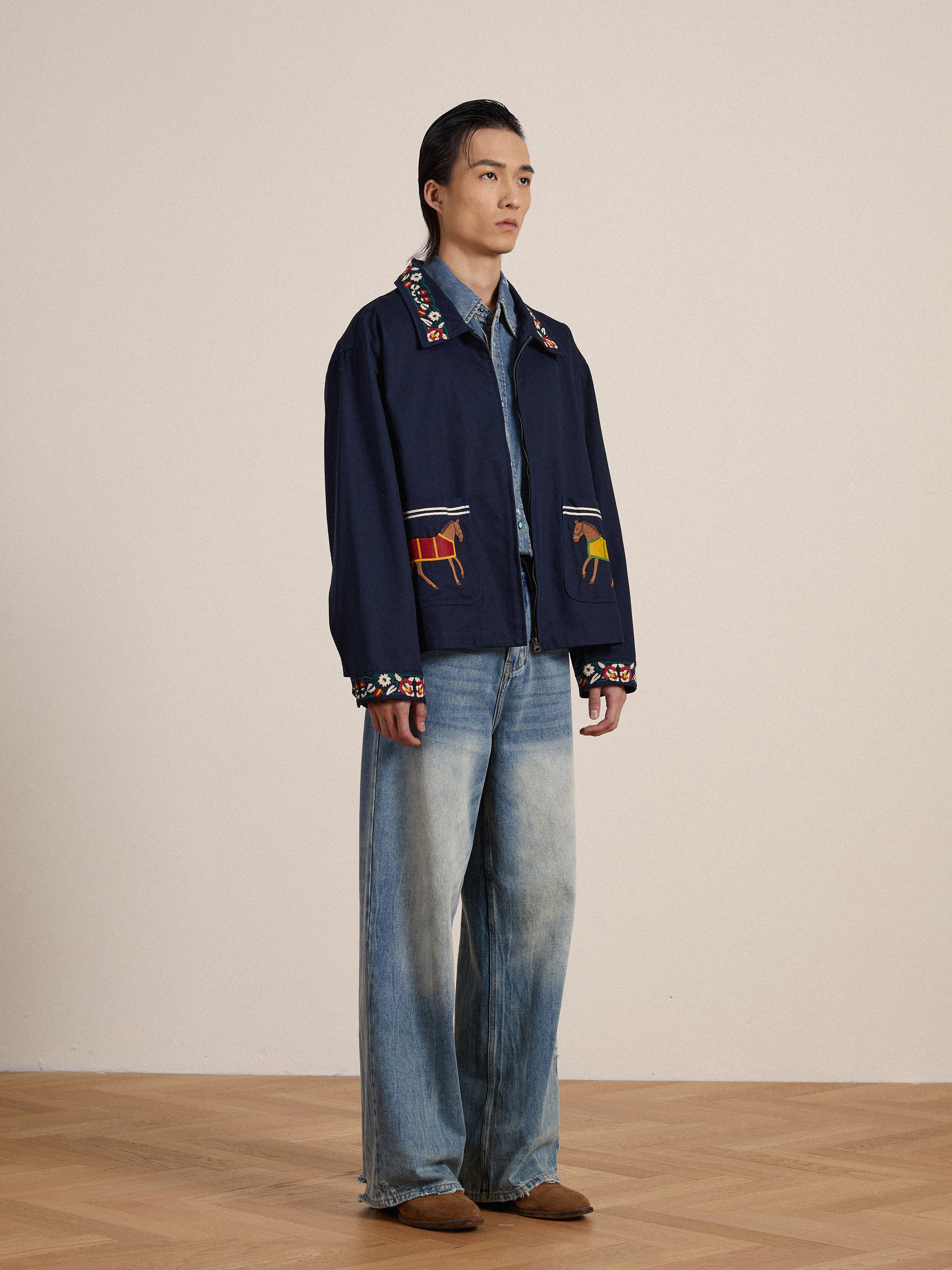 A young man standing in a neutral pose, wearing a navy Found Horse Equine Work Jacket with detailed embroidery and faded jeans against a beige background.