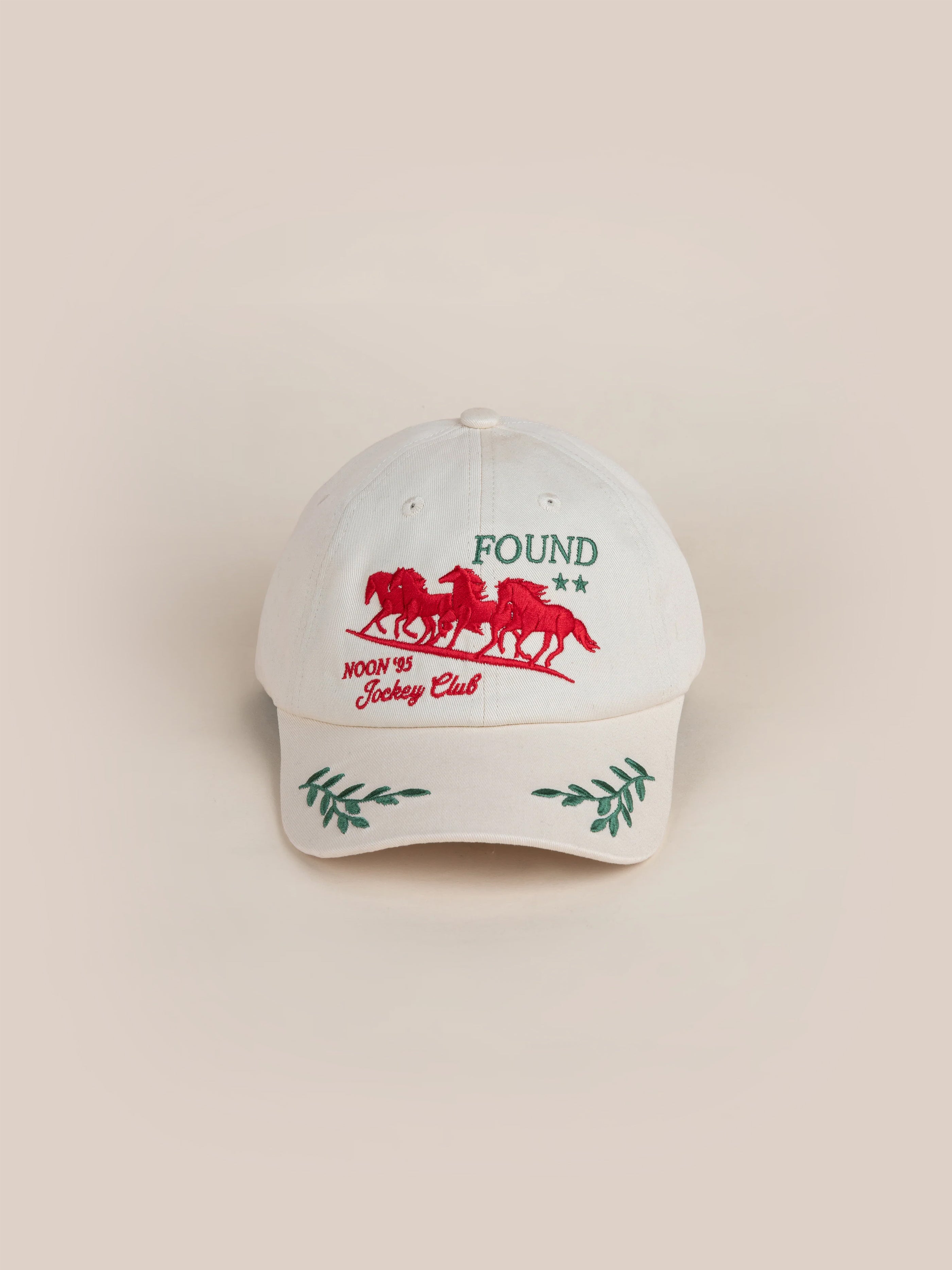 The FOUND Jockey Club Found Cap boasts a stylish white design with a red embroidered horse, beautifully encapsulating the spirit of galloping horses. Adorned with "FOUND Moon J's Jockey Club" text, the cap is further enhanced by vibrant green leaf details on the brim.