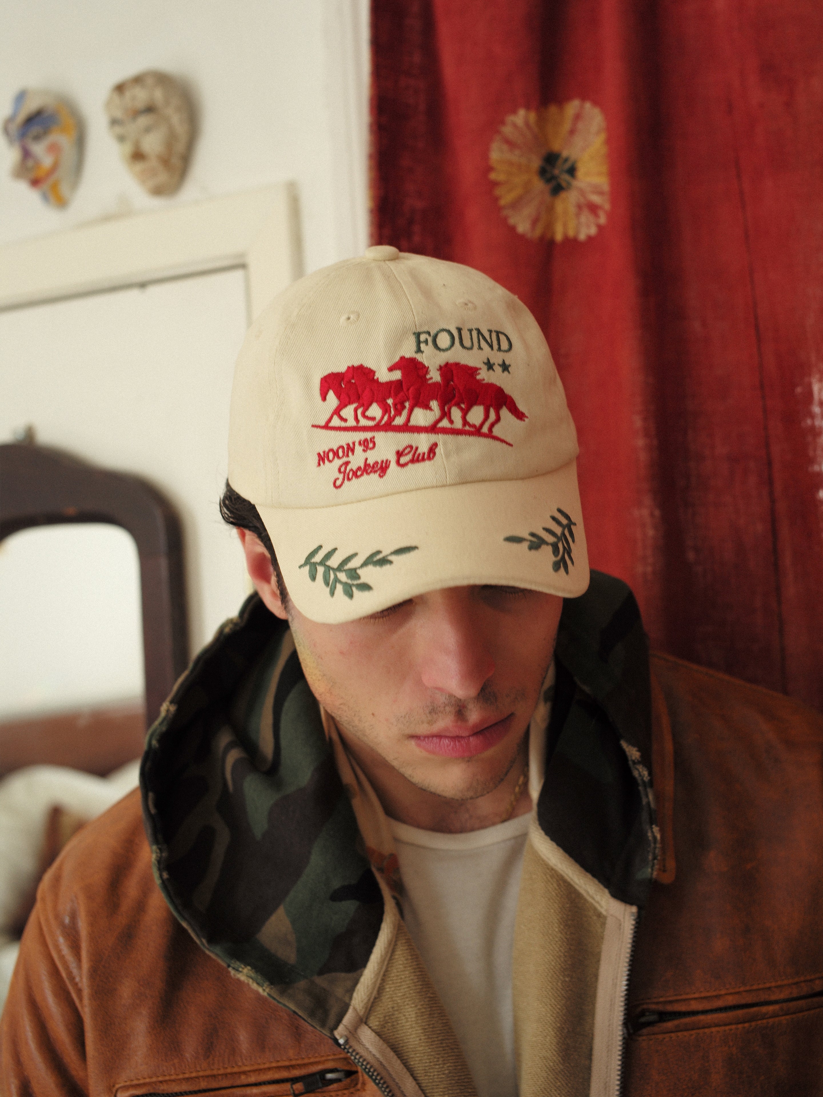 A person in a beige FOUND Jockey Club Found Cap with a horse logo and "NOON 45 Jockey Club," styled with a brown jacket and camo hoodie, stands against a red curtain backdrop adorned with wall decorations.