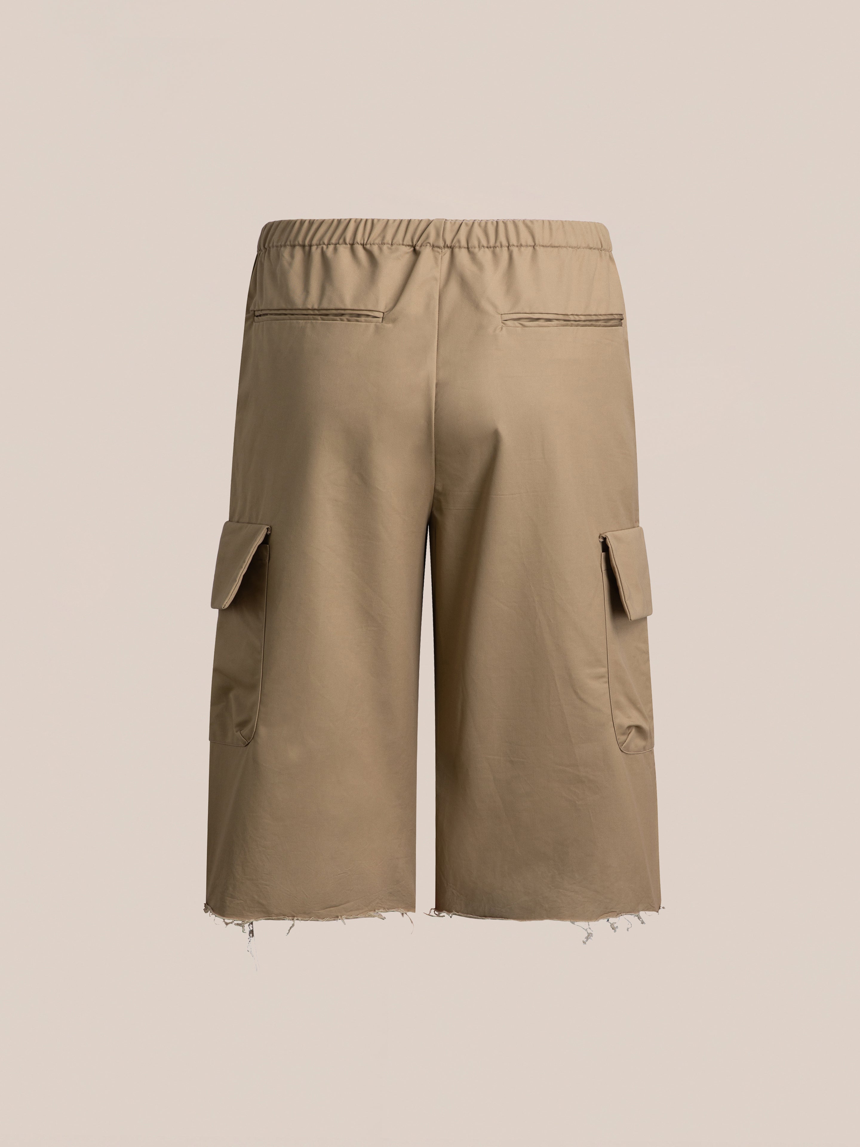 Image of **FOUND Long Twill Cargo Shorts** in beige, featuring an elastic waistband, dual cargo pockets, two back pockets, and two side pockets. The hem is frayed for a distressed look. Made from lightweight cotton fabric. The background is plain light beige.