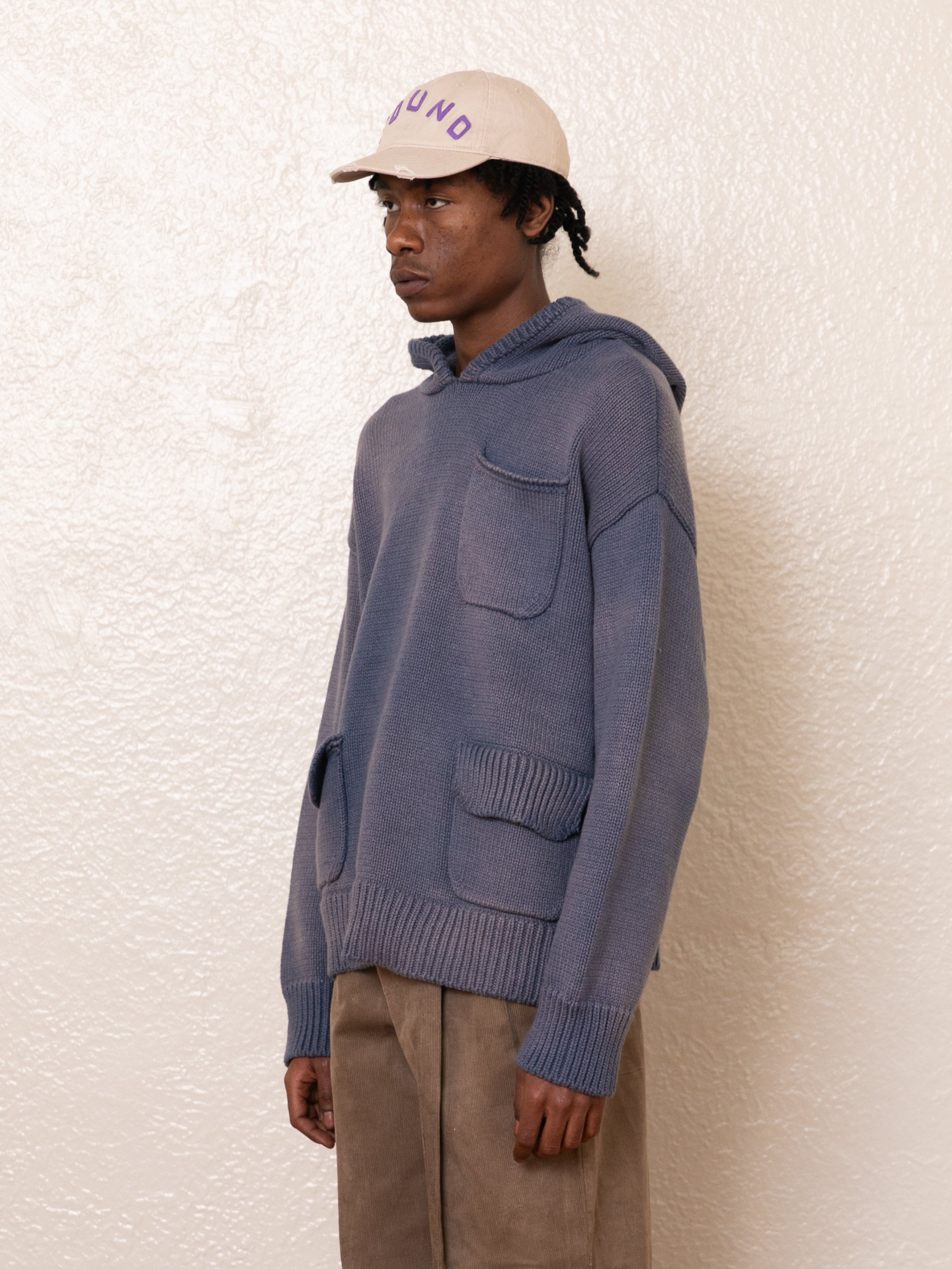 A person in a relaxed oversized beige cap and a FOUND Faded Knit Pocket Hoodie, teamed with 100% cotton brown pants, stands against a textured white wall.