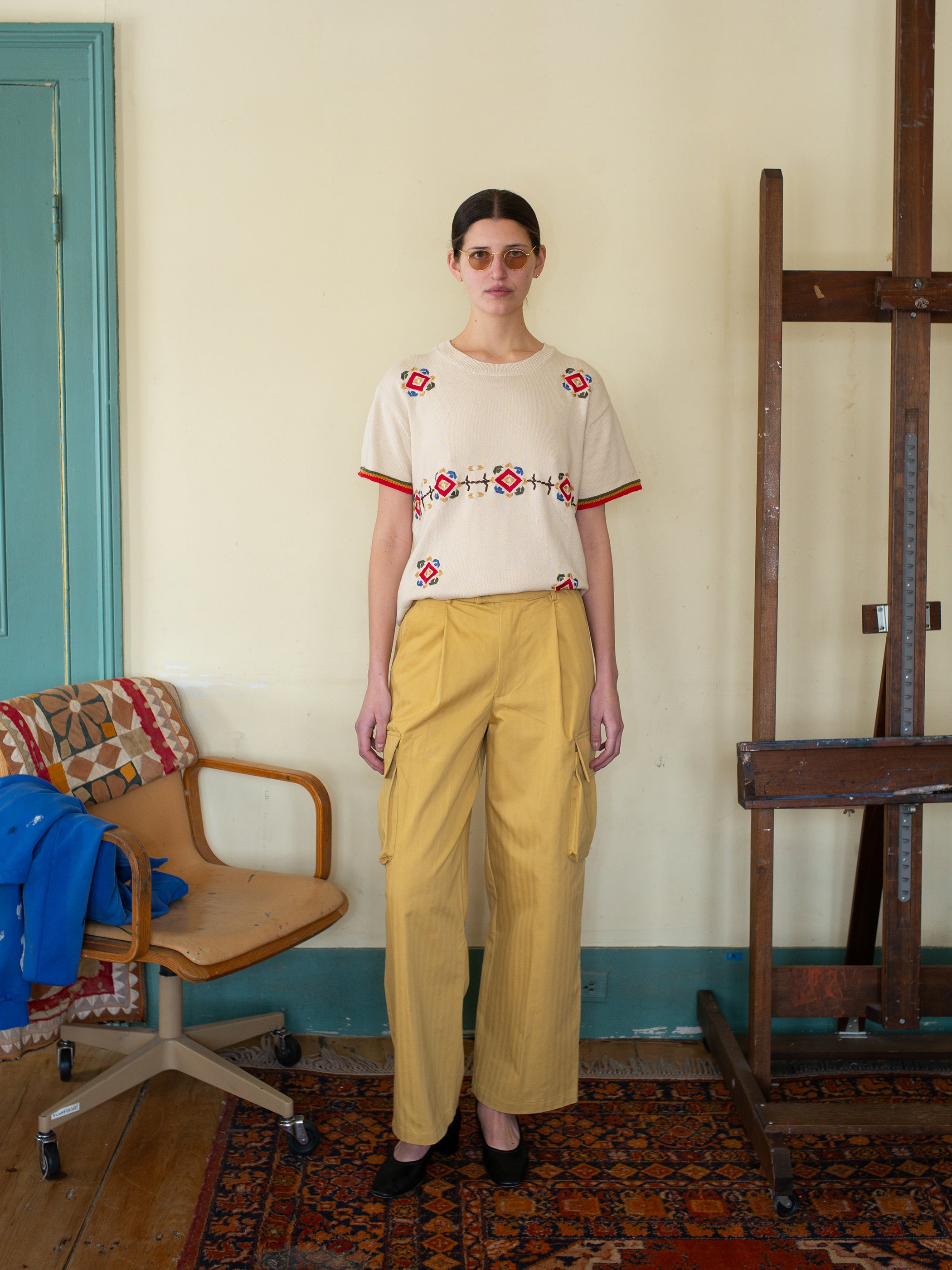 A person wearing the Knit Flower Petal Tee by FOUND with vintage flair stands indoors between a chair and wooden structure on a patterned rug.