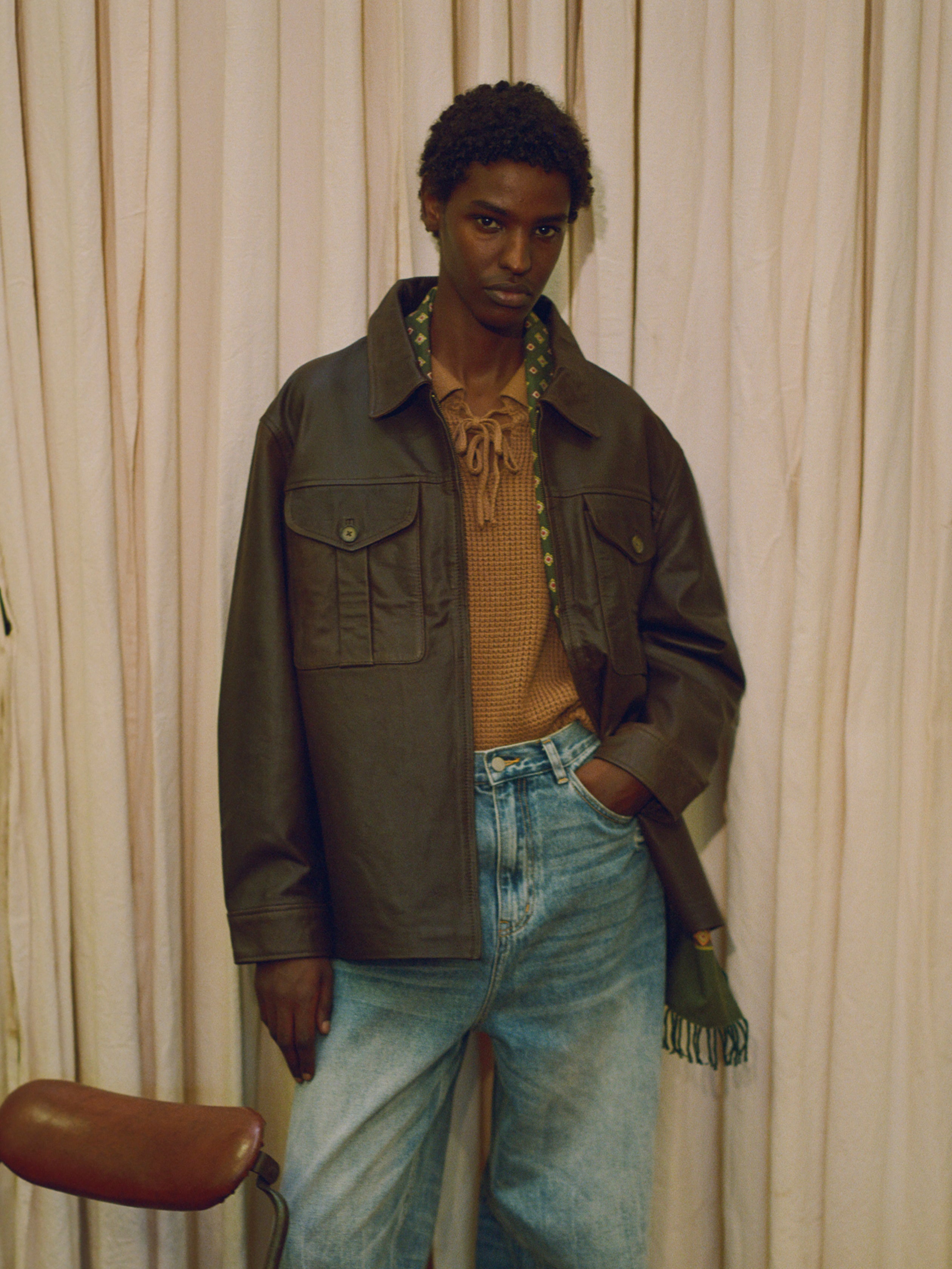 A man in a cozy brown jacket and jeans leaning against a curtain, enjoying the autumn vibes in his Found Tie-Collar Knit Sweater.