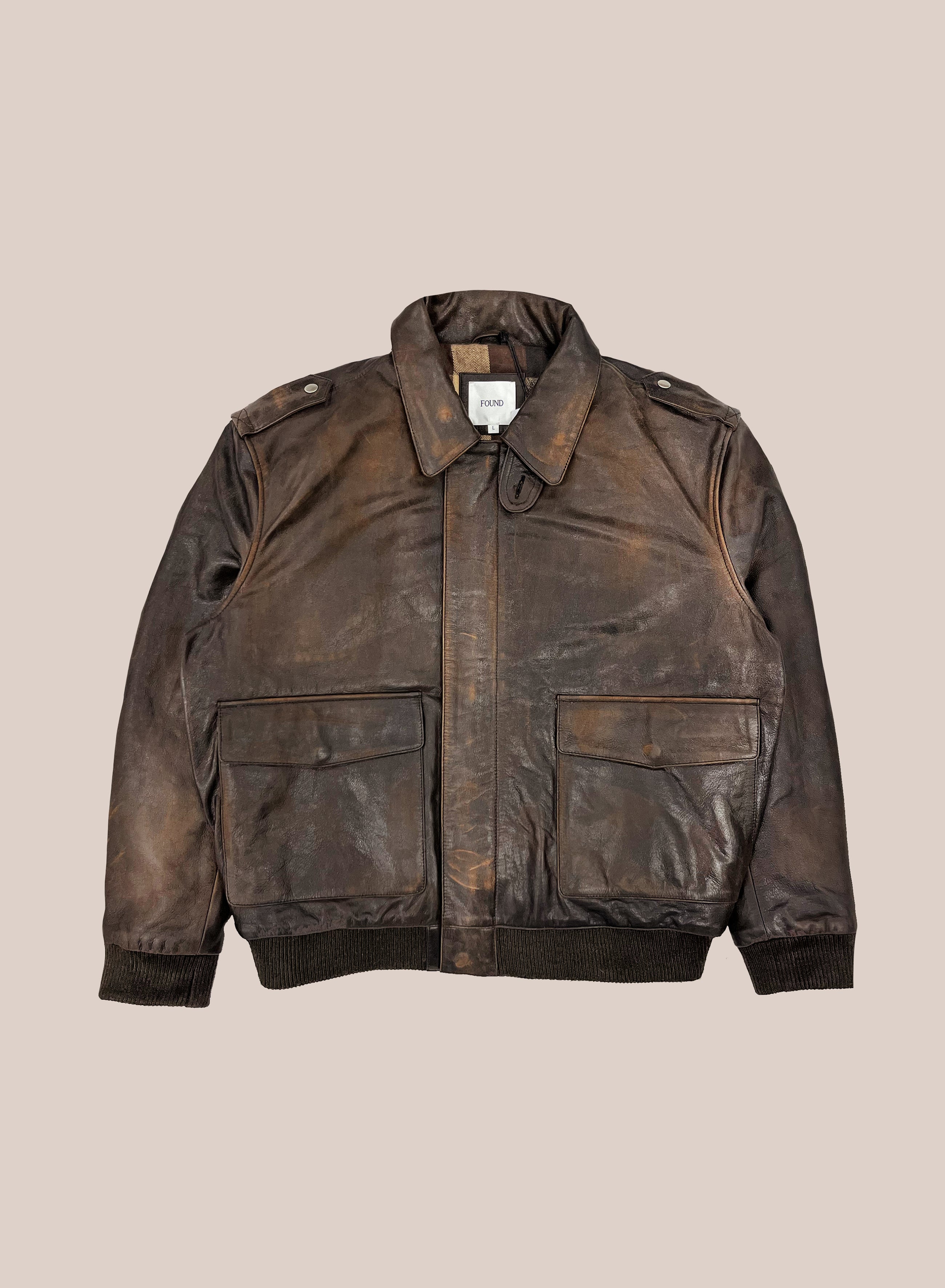 The FOUND Distressed Leather Flight Jacket features a brown distressed finish, front flap pockets, a collar, and ribbed cuffs and hem, shown against a plain beige background.