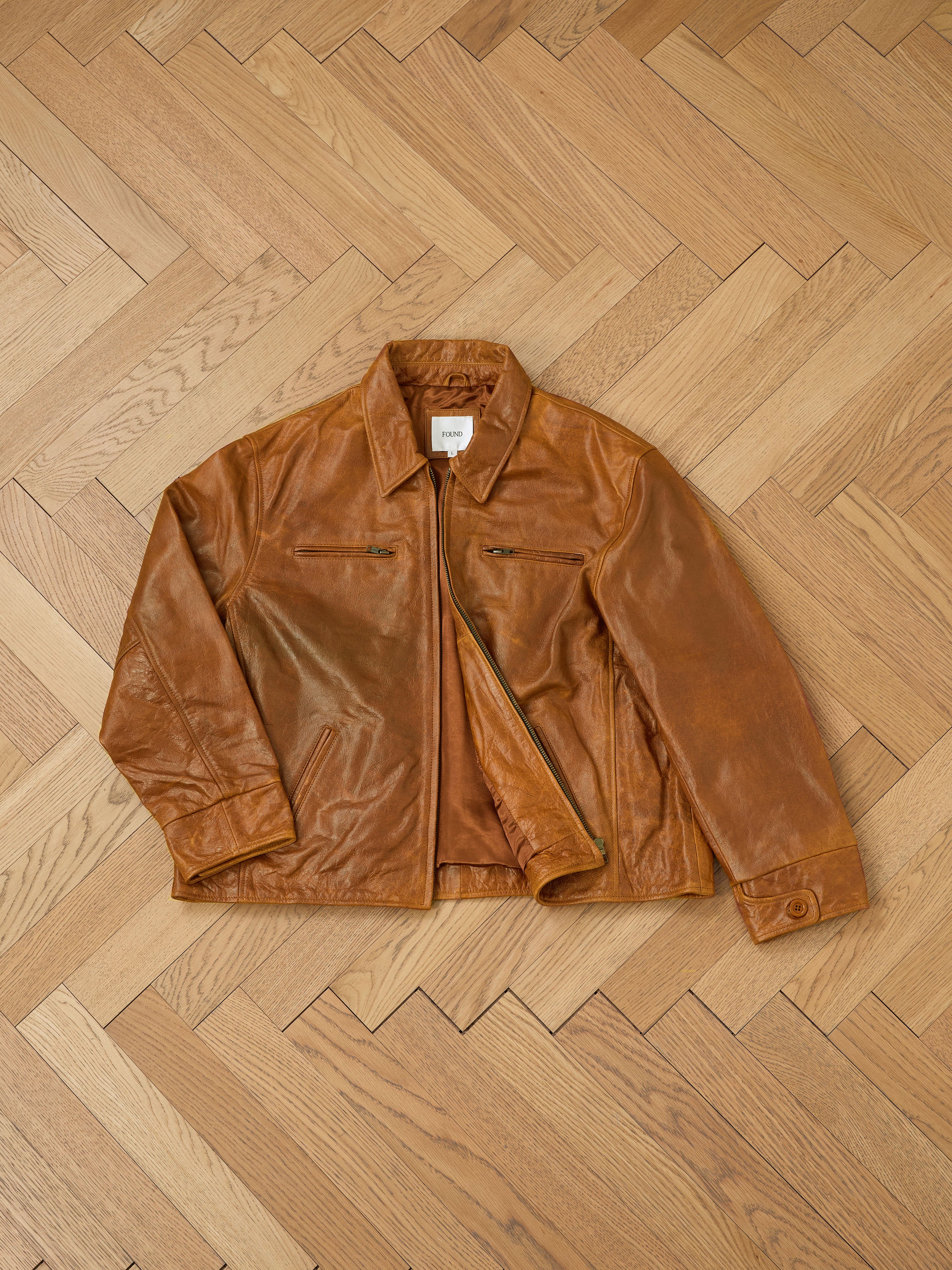 The Marlboro Sun Faded Leather Racer Jacket by FOUND, a brown leather piece with a zipper, made from ethically sourced materials, is laid flat on a wooden herringbone-pattern floor.