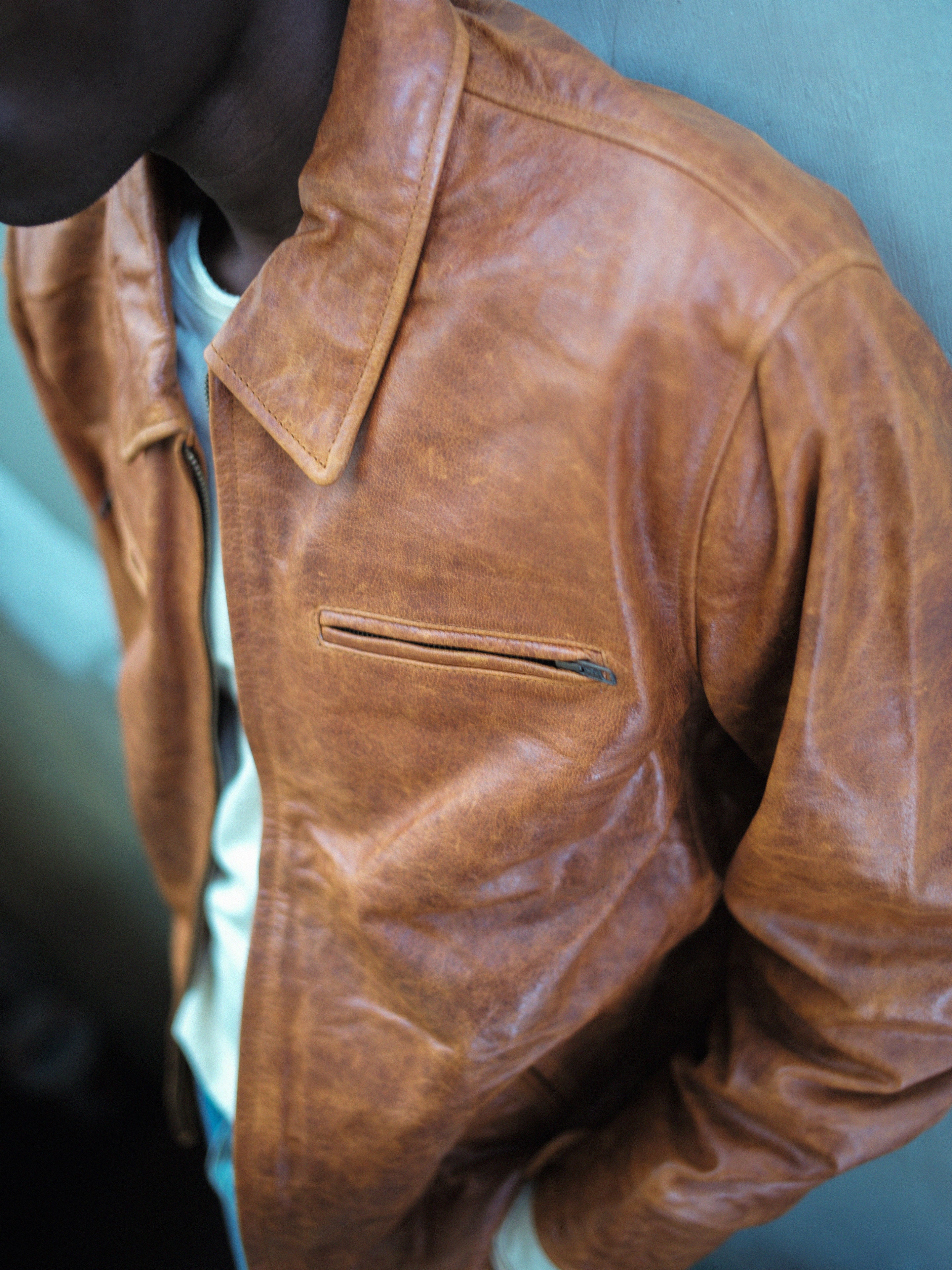 Donning the FOUND Marlboro Sun Faded Leather Racer Jacket with its signature front zipper pocket, they stand confidently against the wall, showcasing timeless style crafted from ethically sourced materials.