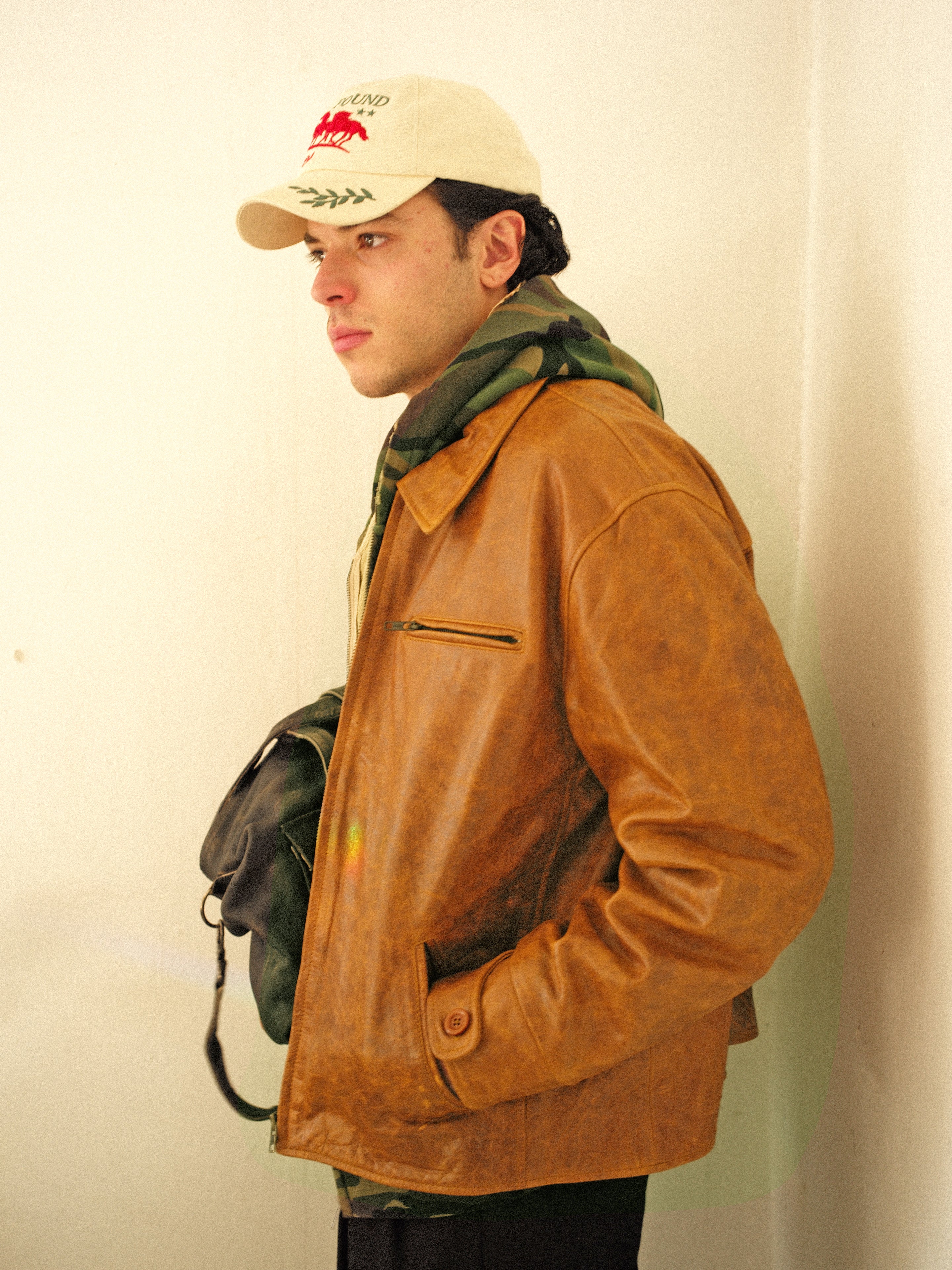 In profile view, a person exudes casual sophistication by pairing a beige cap and green scarf with the Marlboro Sun Faded Leather Racer Jacket from FOUND. They hold a black bag against a plain white wall.