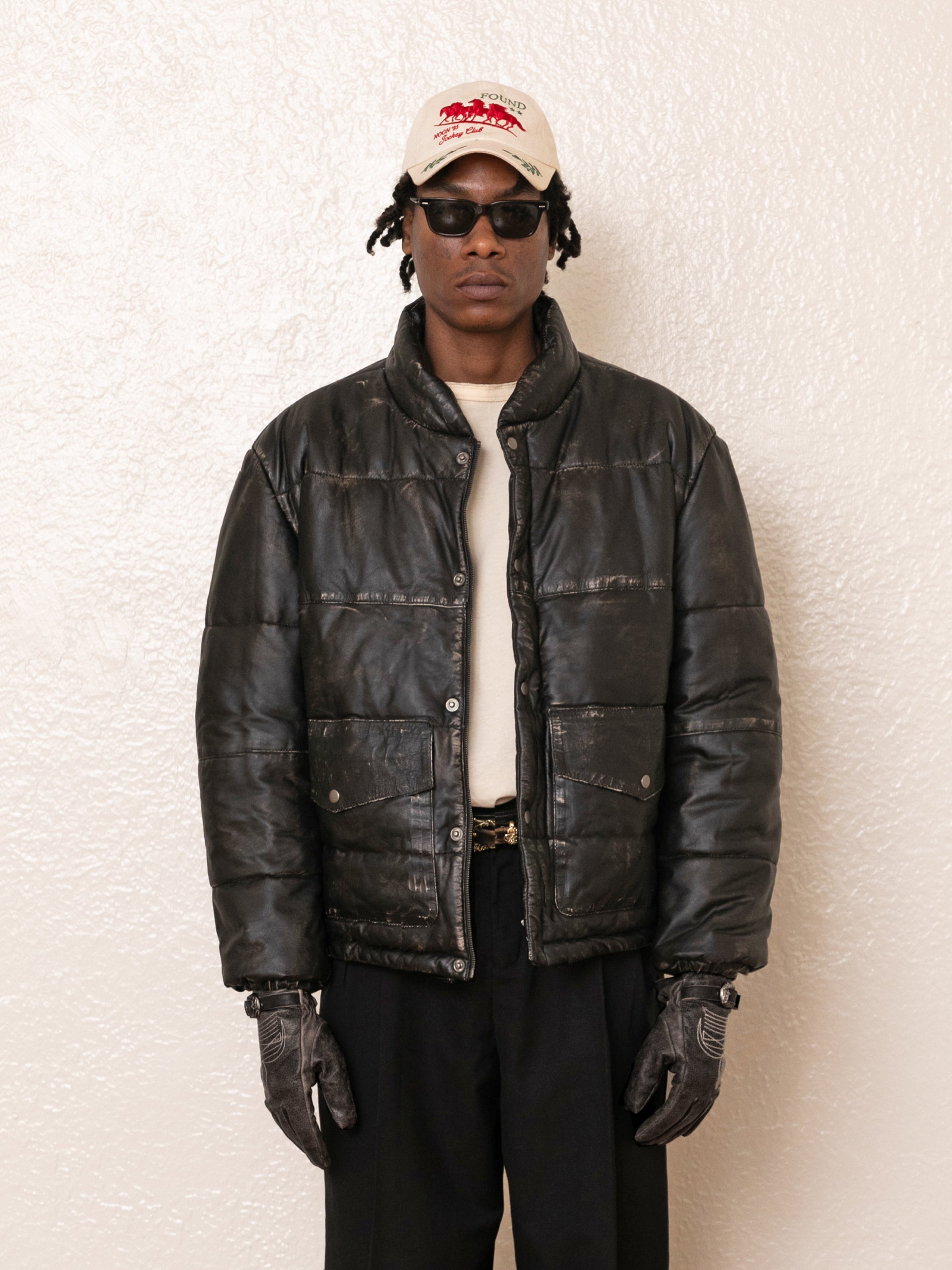 A person in a FOUND Leather Distressed Puffer Jacket, gloves, sunglasses, and a white cap stands against a textured white wall.