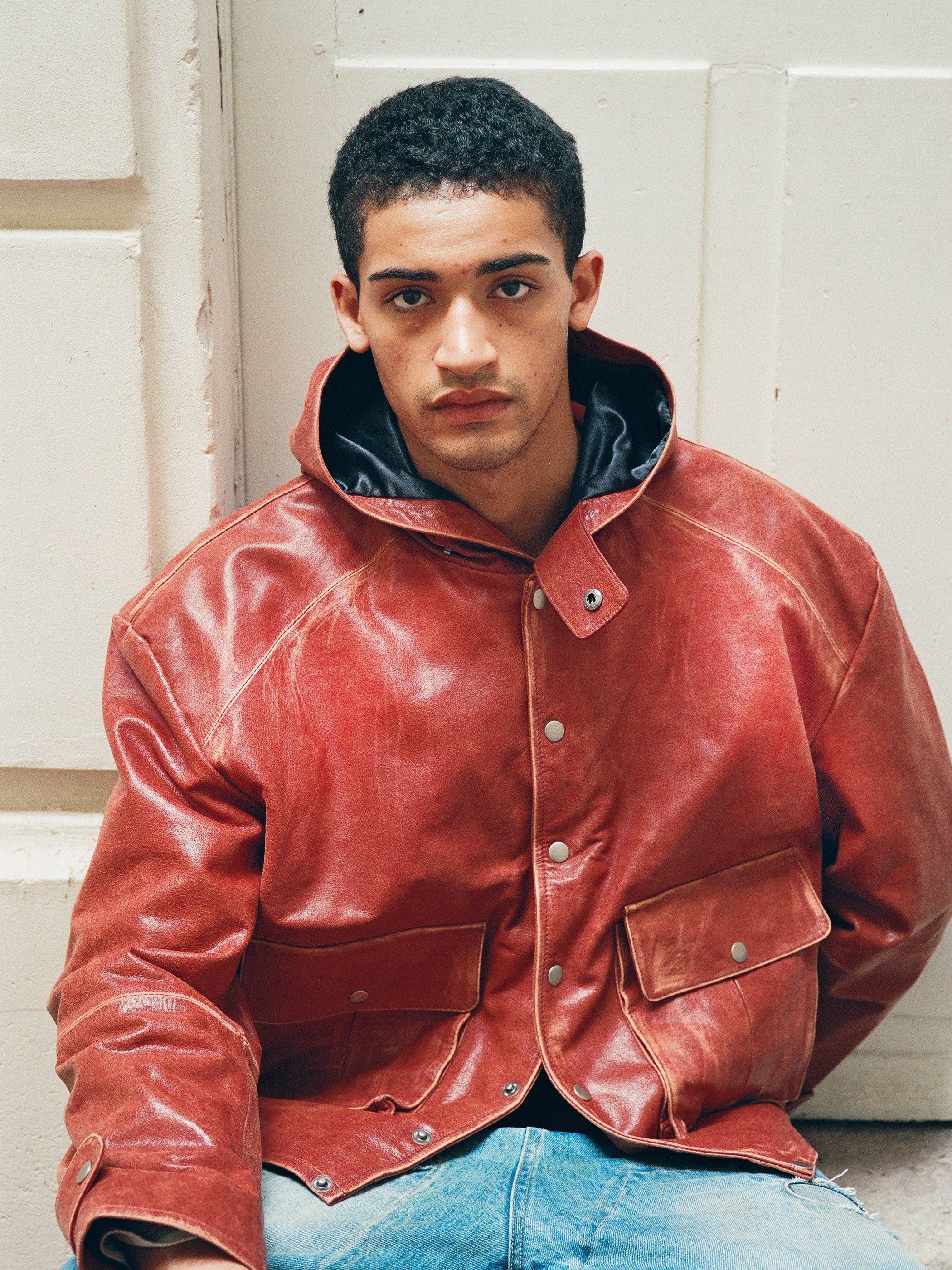 A person with short curly hair wears the FOUND Distressed Leather Pocket Hoodie in vintage red and light blue jeans, sitting against a white wall and looking at the camera.
