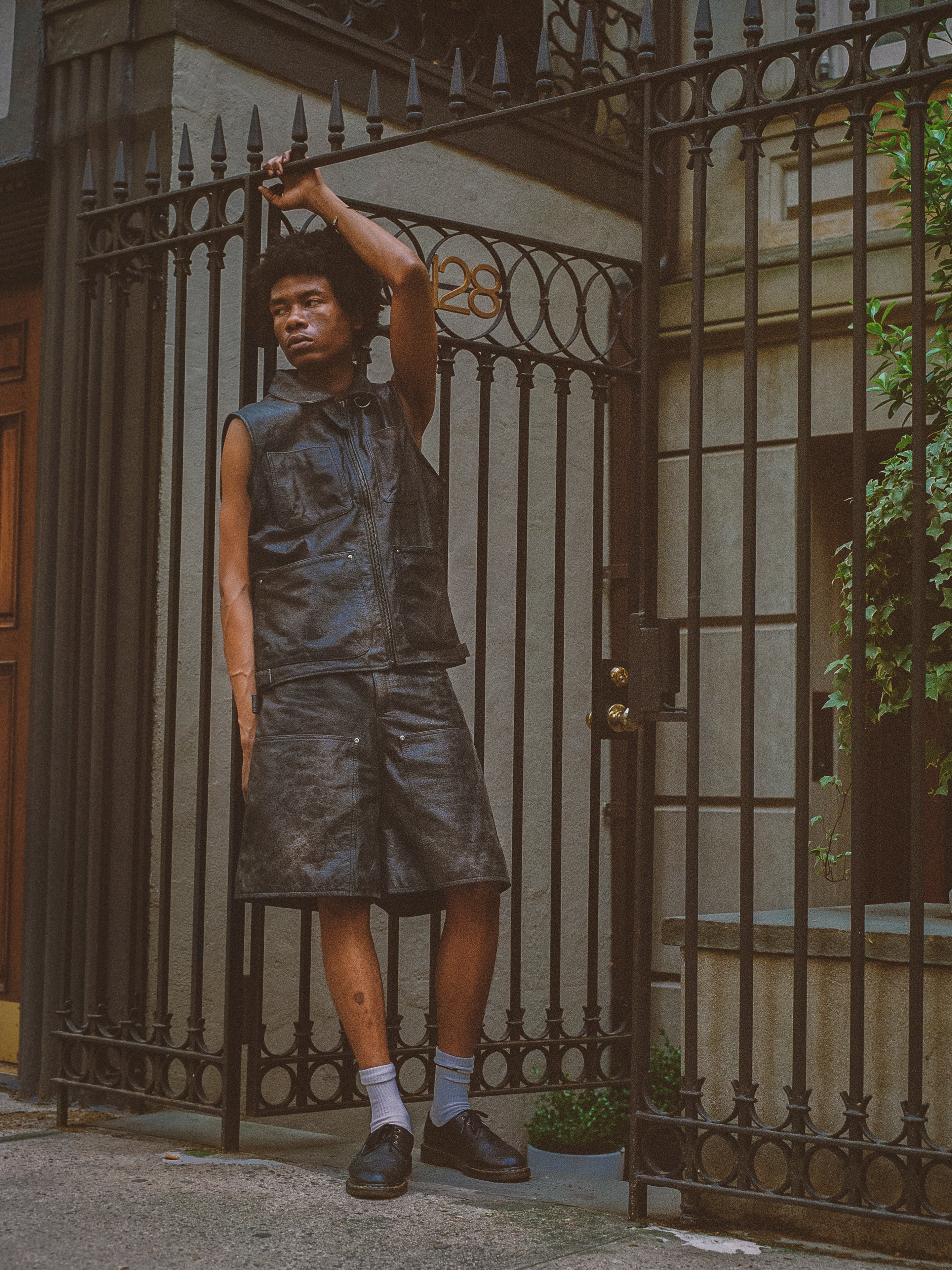 A person in a Found Distressed Genuine Leather Pocket Vest leans on a wrought iron fence, with a building in the background.