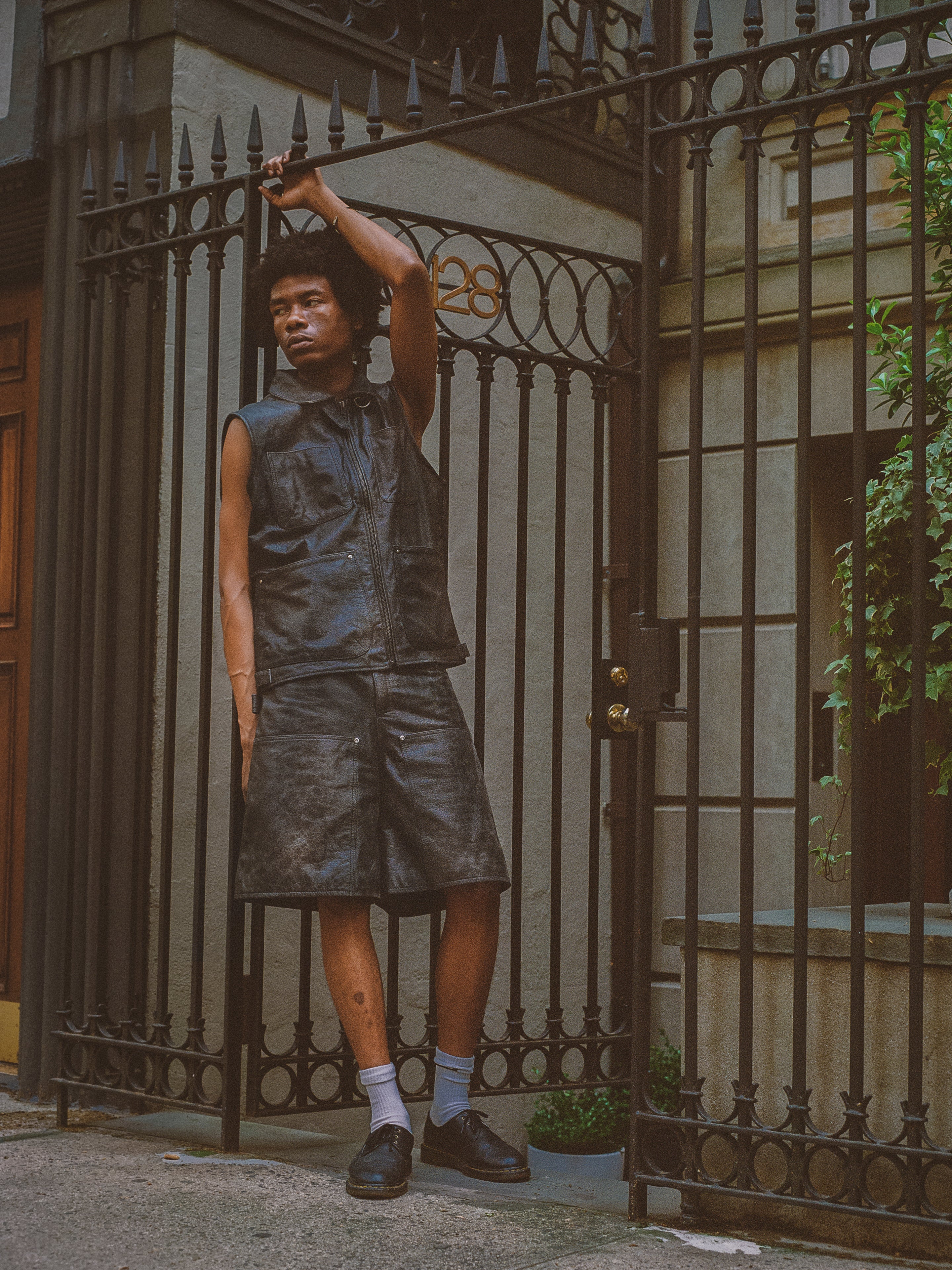 A person stands in front of a wrought iron gate, wearing a sleeveless leather vest and Found Distressed Genuine Leather Carpenter Shorts with a double-knee silhouette. One arm is raised, holding onto the gate, which has the address number "28" above it.