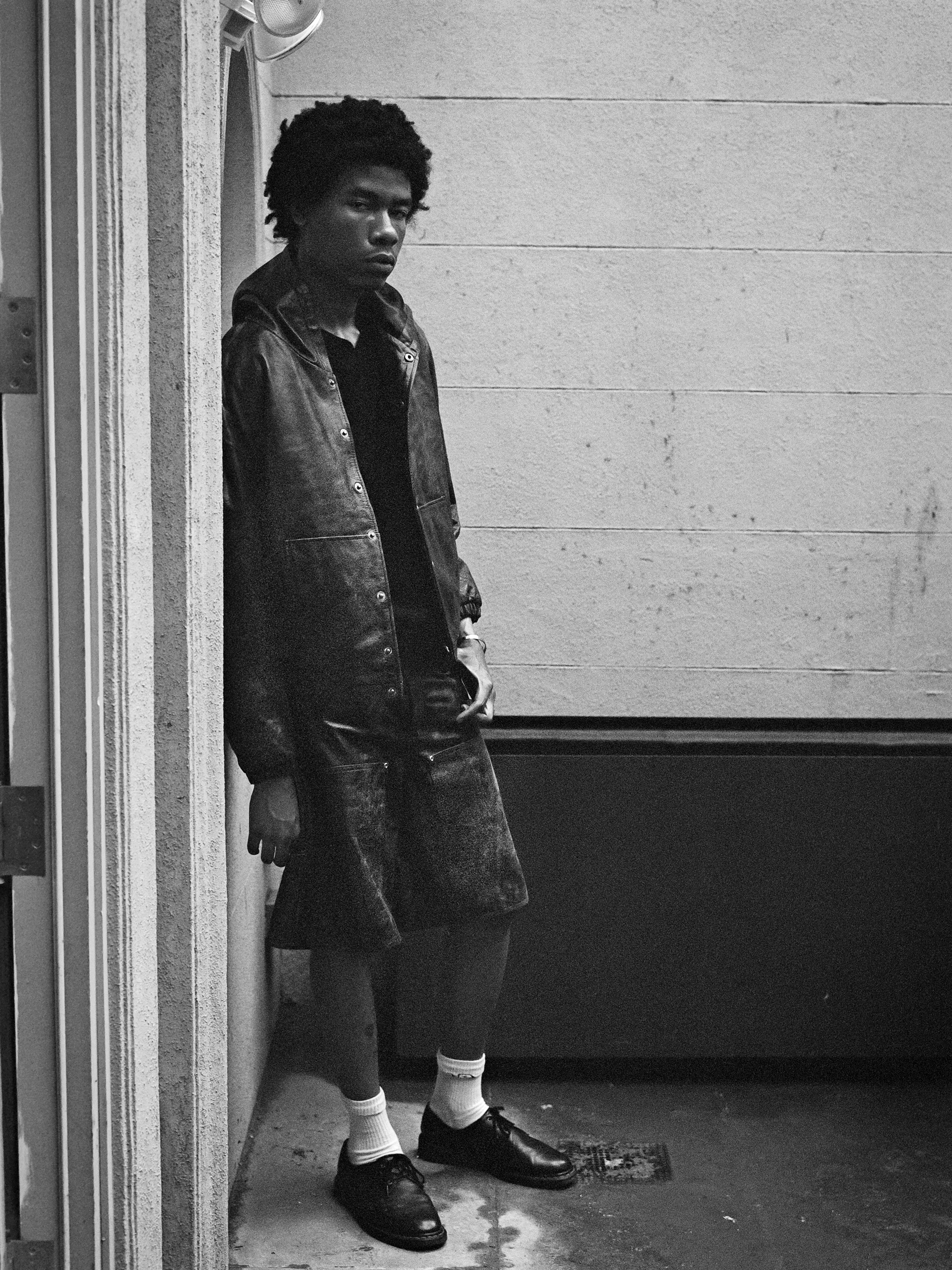 A person with short, curly hair stands in a narrow corridor, wearing the Distressed Genuine Leather Hoodie by Found, along with boots, shorts, and socks. They gaze off to the side in this black and white photograph.