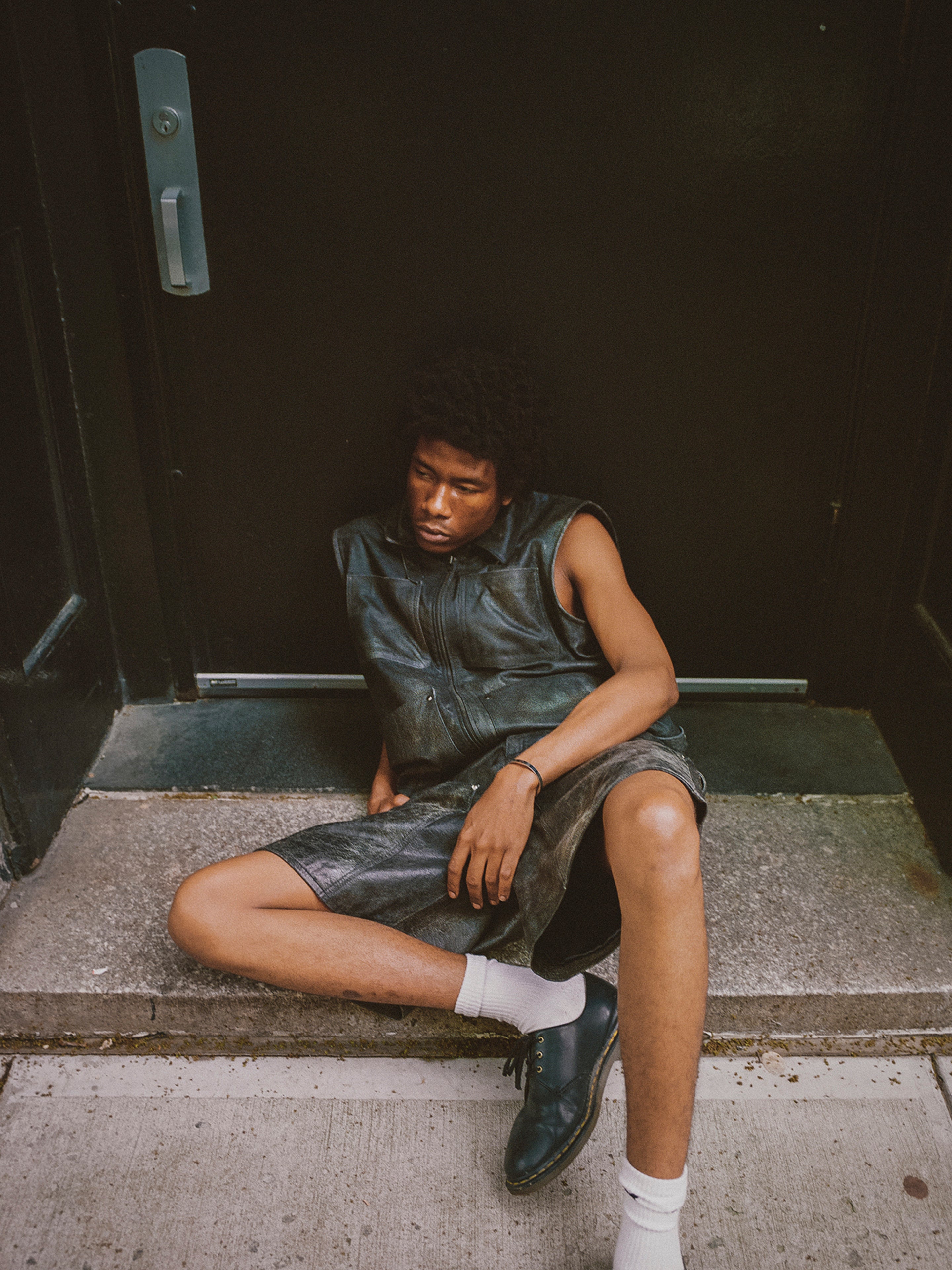 A person with short curly hair sits on a doorstep, donning a Found Distressed Genuine Leather Pocket Vest, matching shorts, white socks, and black shoes, looking downward.