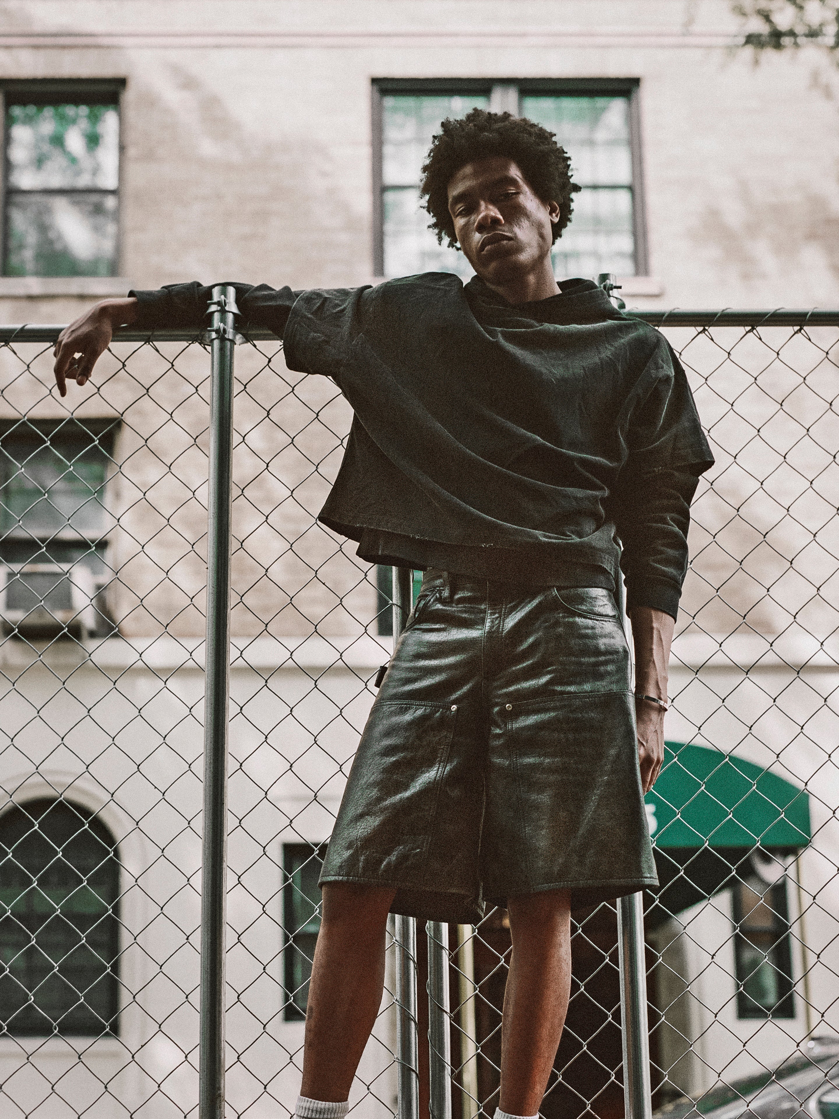 A person in dark clothing, featuring Found's Distressed Genuine Leather Carpenter Shorts, leans against a chain-link fence in an urban area with buildings in the background.