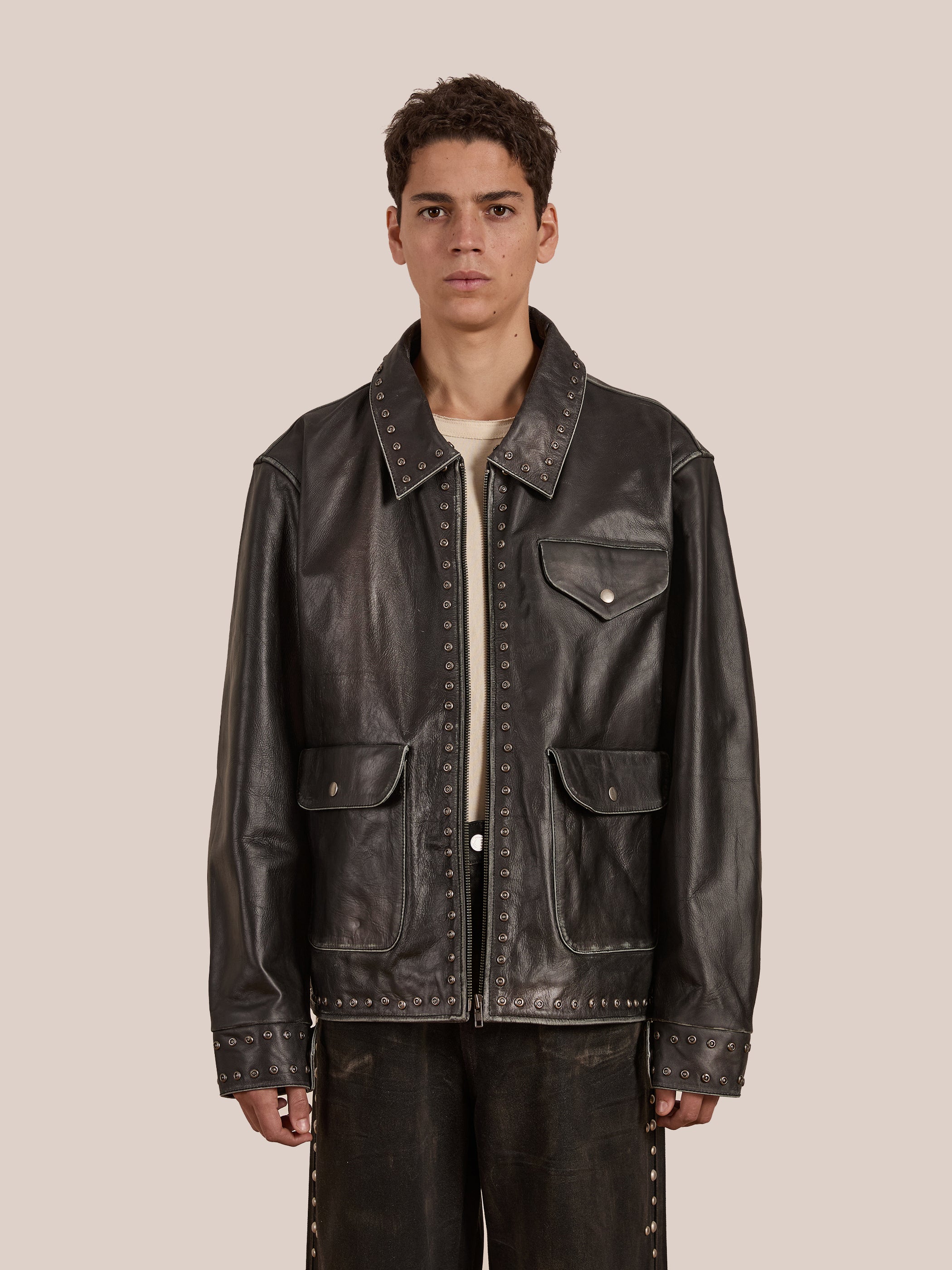 A person in FOUND's Studded Leather Pocket Jacket, against a plain beige background, styles it over a light shirt for a timeless look.