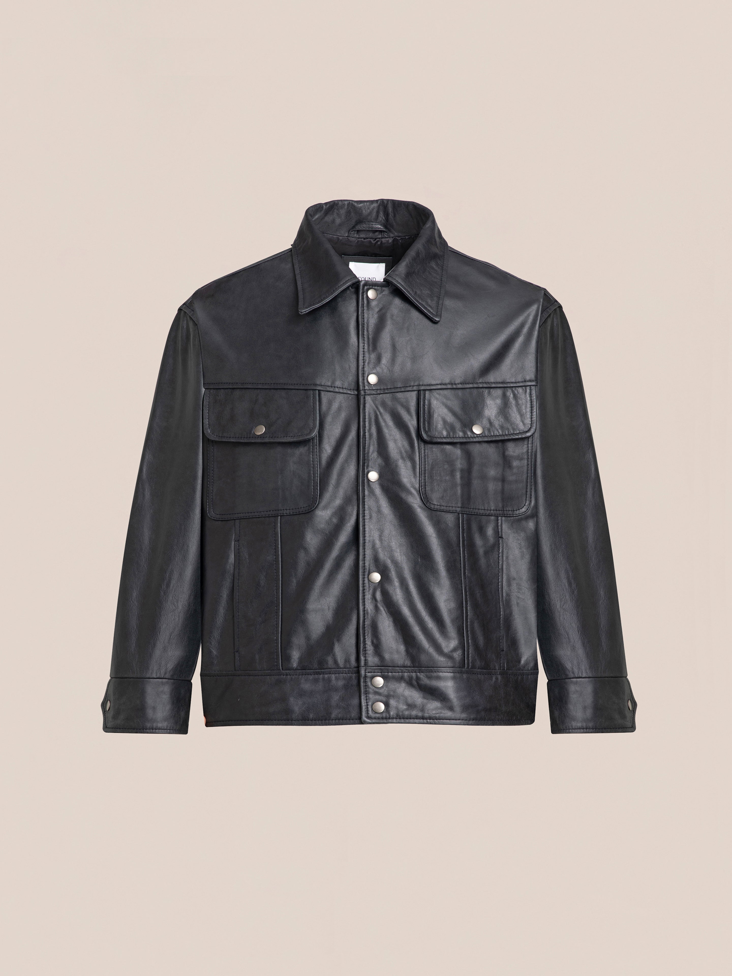 The Leather Trucker Jacket by FOUND features a classic black leather design with a button-up front, two chest pockets with flaps, and a collar, showcased against a beige background.