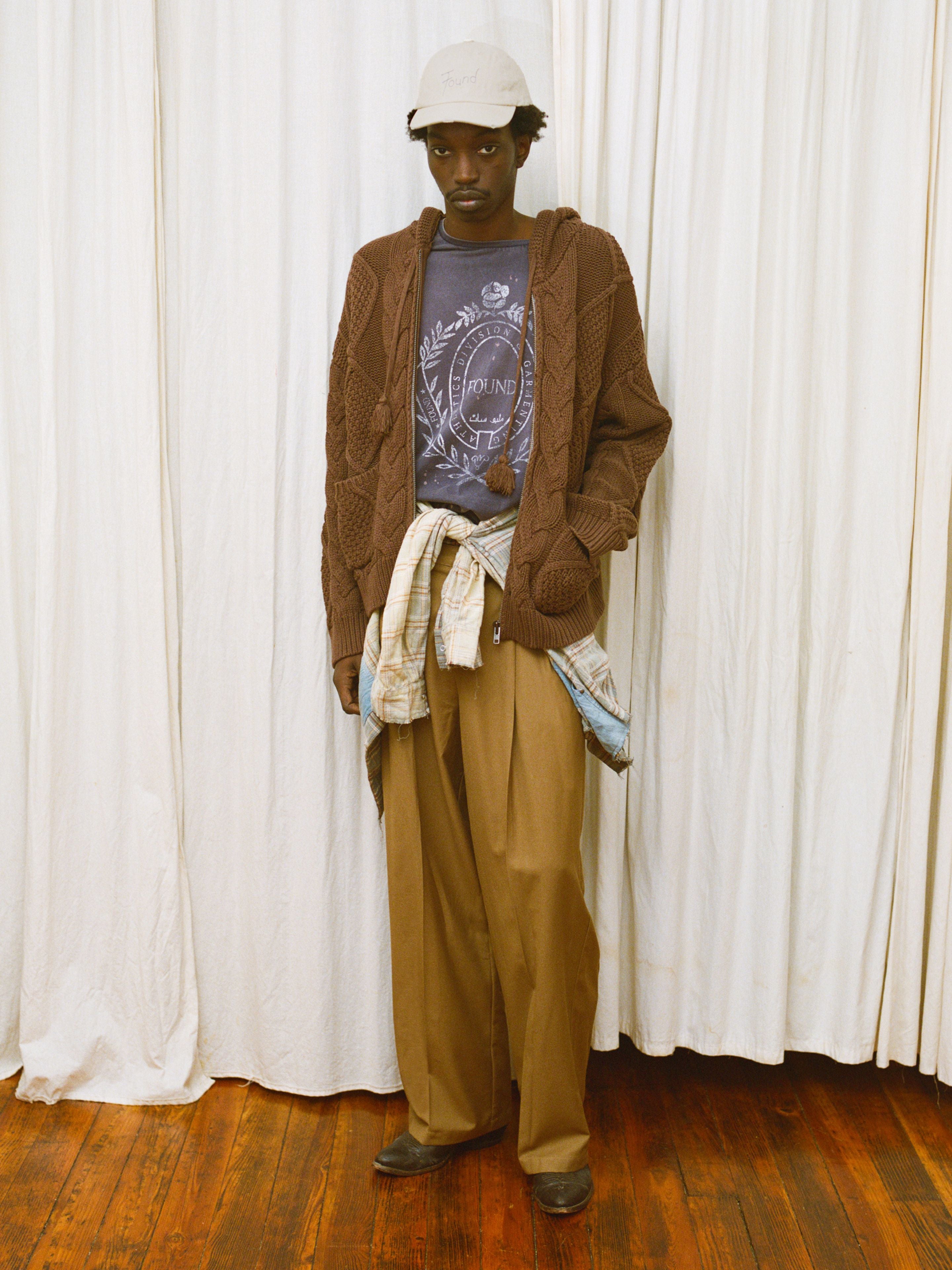 A person stands indoors in a beige cap, brown cardigan, FOUND Collegiate Crest Tee, tan pants, and a plaid shirt tied at the waist, in front of white curtains on a wooden floor.