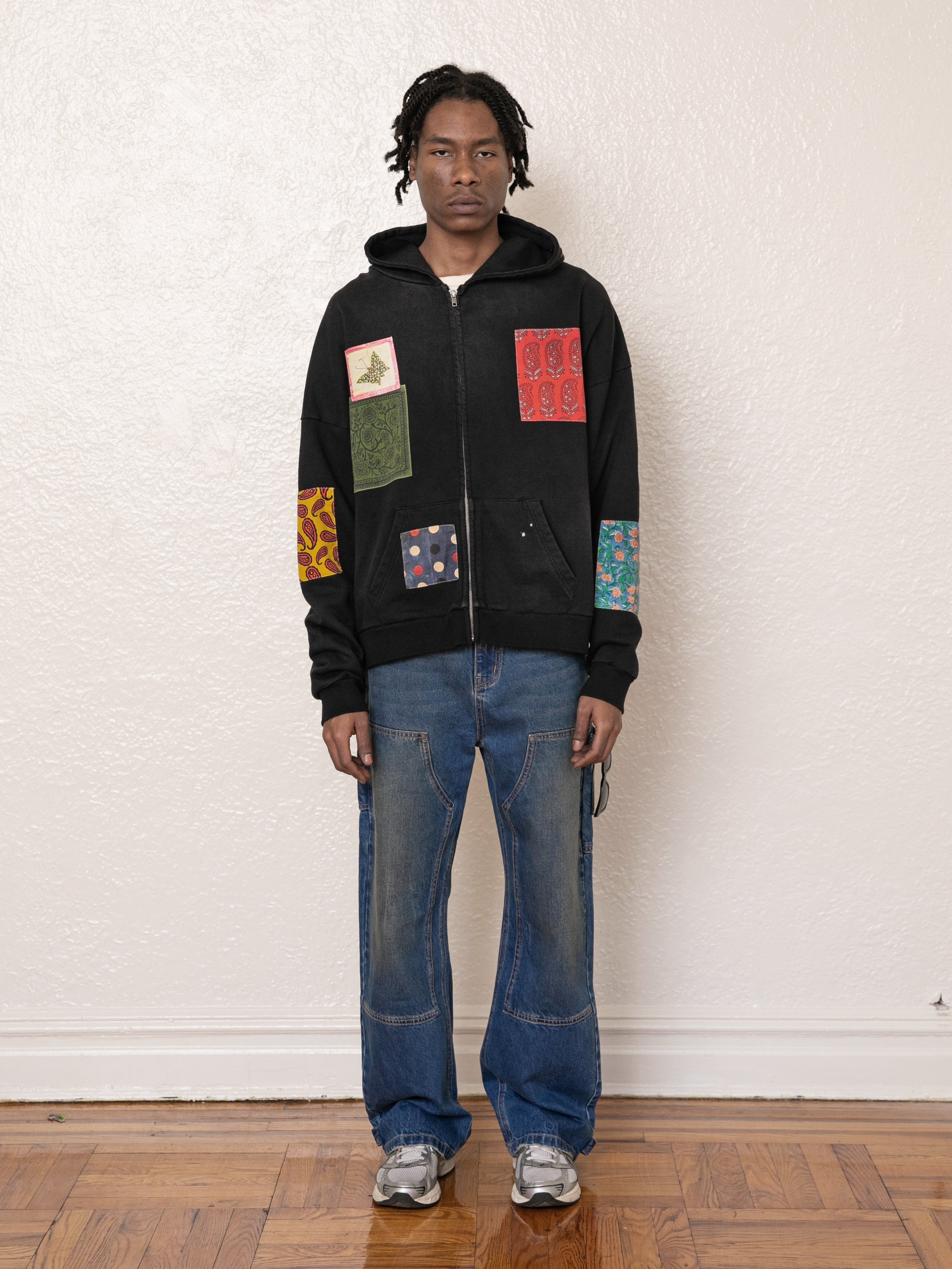 A person wearing a FOUND Tapestry Patchwork Zip Up Hoodie with blue jeans and sneakers stands against a plain wall.