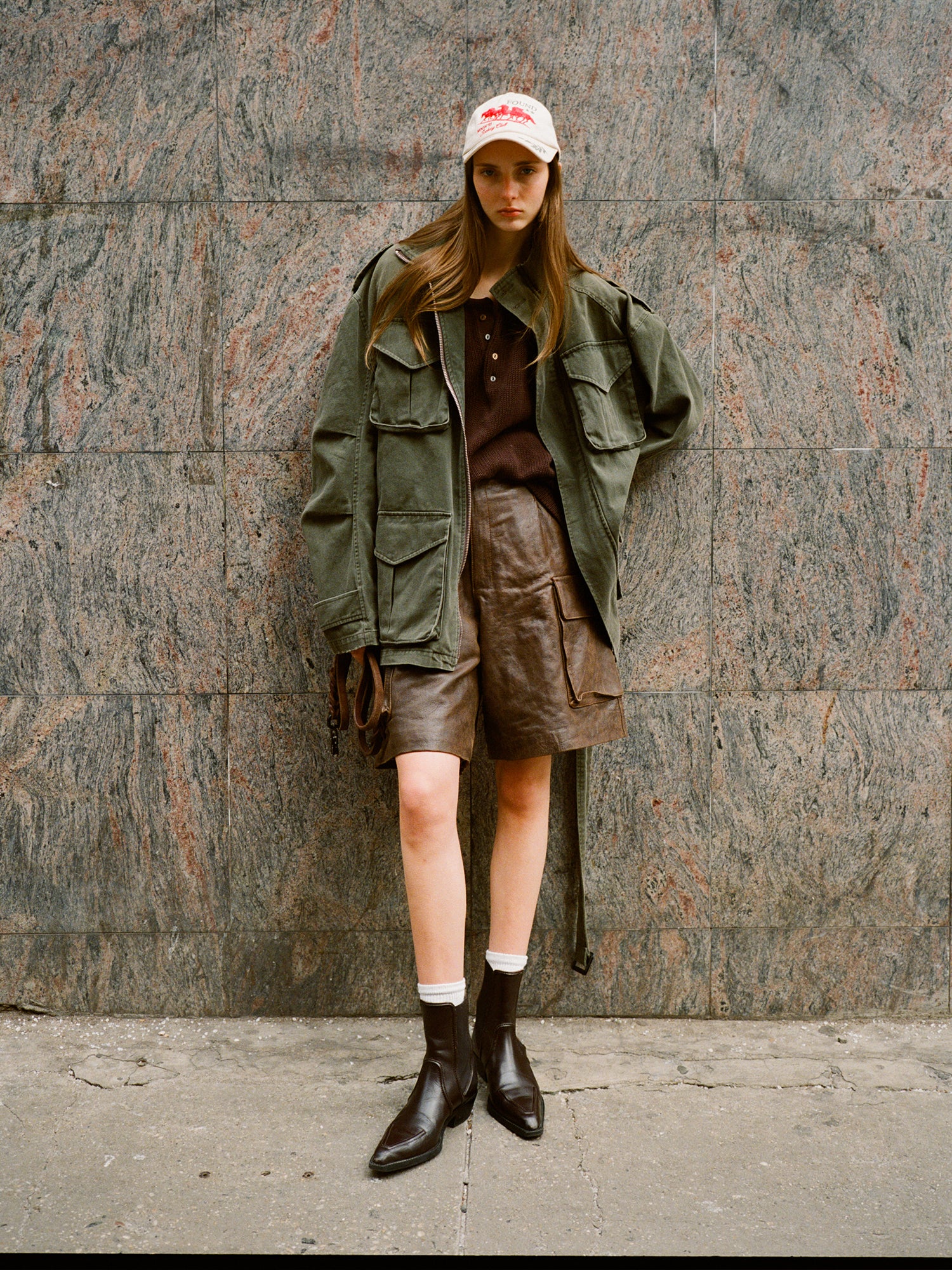 A person in an oversized FOUND M65 Utility Twill Pocket Jacket, leather shorts, and boots poses against a textured wall, sporting a cap. The military-inspired outerwear enhances the vintage look for a bold style statement.