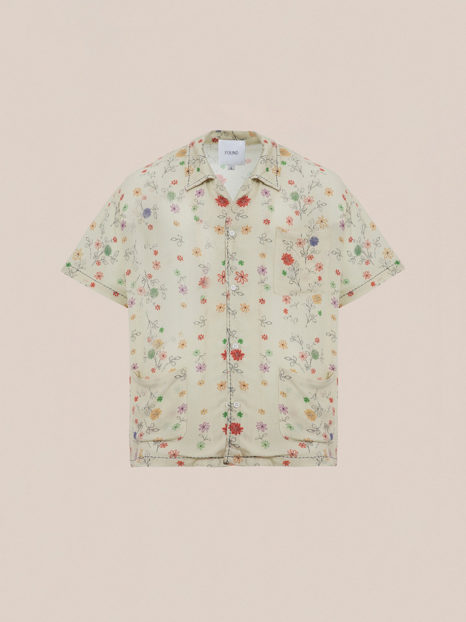 The Profound Multi Floral Camp Shirt is a short-sleeve, button-up made from lightweight rayon fabric, showcasing a light beige background with a vibrant floral print. It features a notched collar, front pockets, and an inner neck label for added detail.