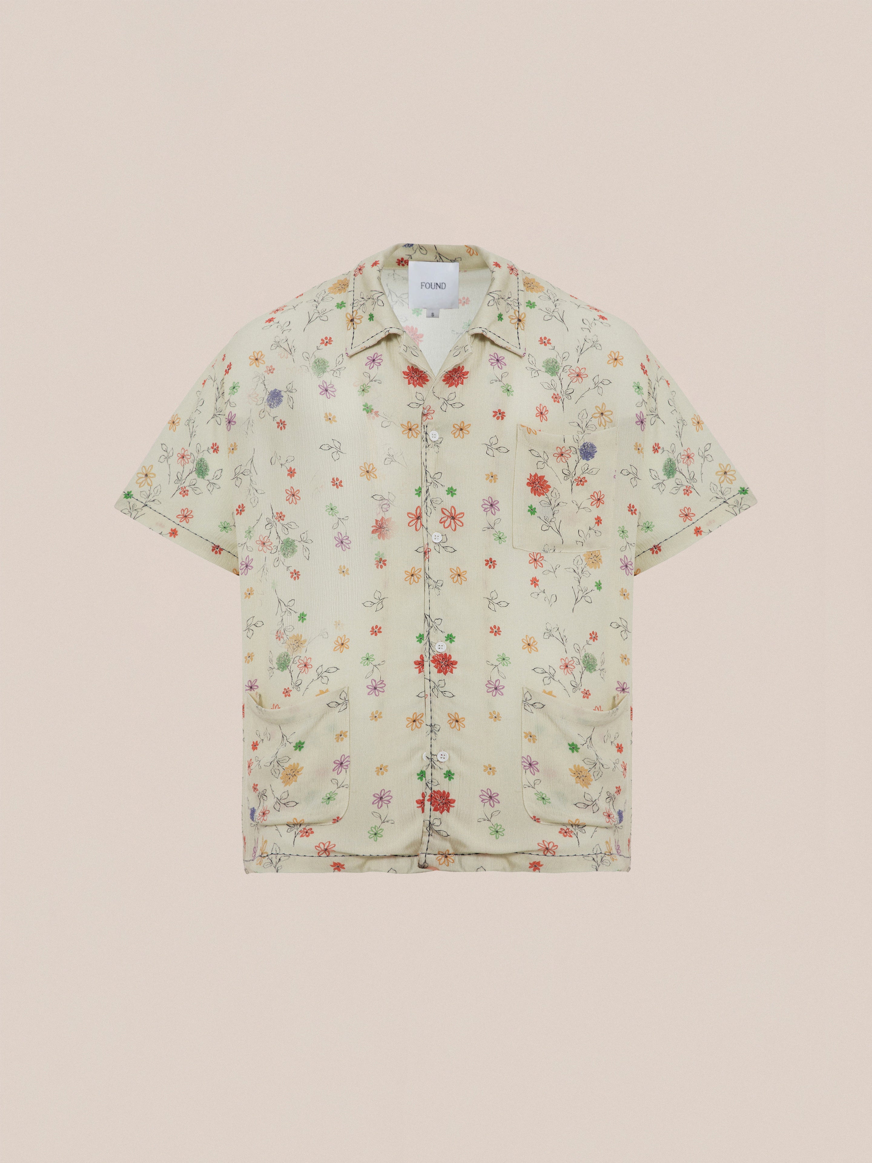 The Profound Multi Floral Camp Shirt is a short-sleeve, button-up made from lightweight rayon fabric, showcasing a light beige background with a vibrant floral print. It features a notched collar, front pockets, and an inner neck label for added detail.