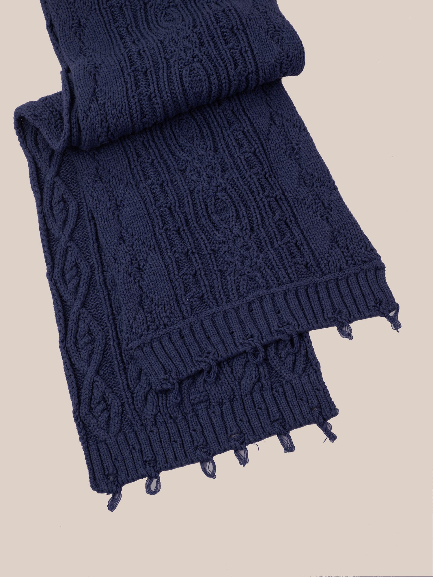 A partially folded FOUND Navy Distressed Cable Knit Scarf with fringe ends rests on a light surface.