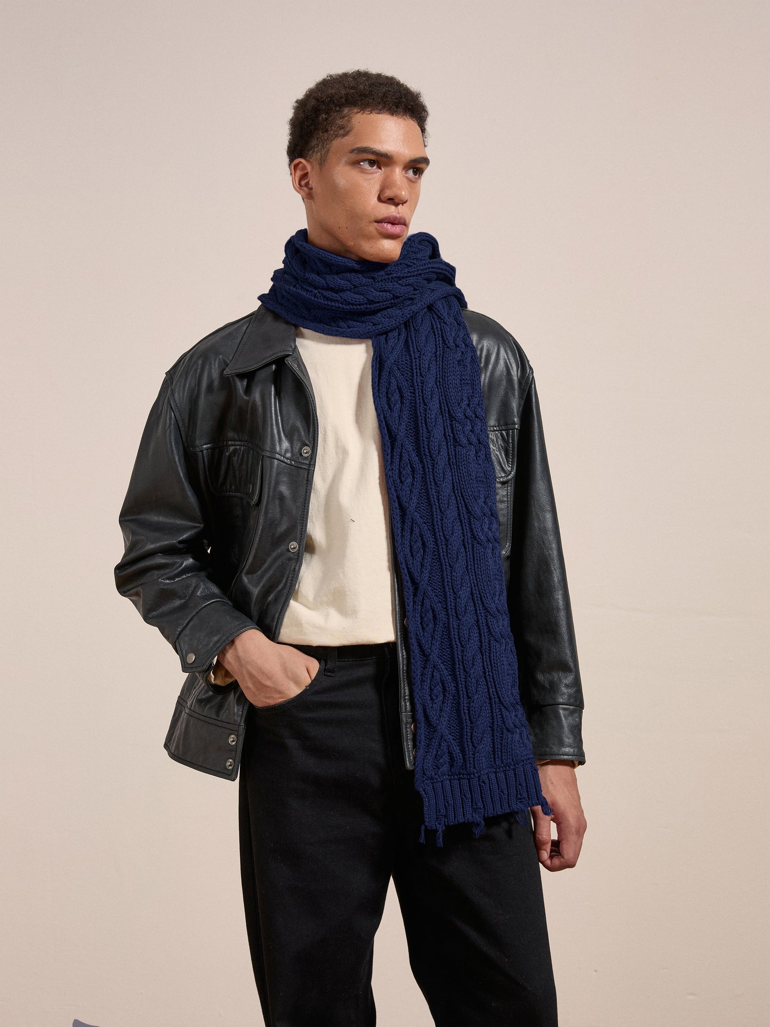 A person with short hair is wearing a leather jacket over a light shirt and a FOUND Navy Distressed Cable Knit Scarf. They are standing against a plain background, looking off to the side.