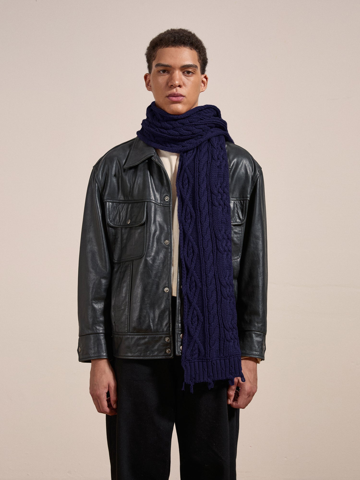 A person stands against a plain background wearing a leather jacket and FOUND's Navy Distressed Cable Knit Scarf.