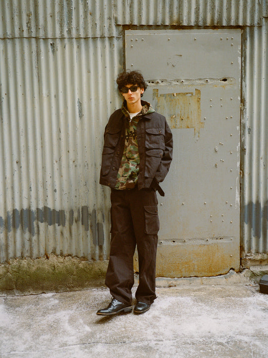 Against a corrugated metal wall with a closed door, someone wearing sunglasses and a vintage-look camo shirt sports the FOUND Utility Herringbone Cargo Jacket.