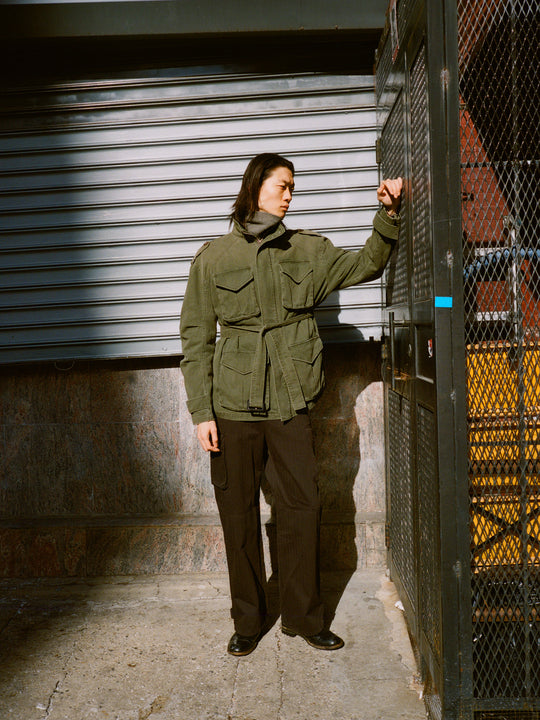 Wearing a vintage style M65 Utility Twill Pocket Jacket by FOUND and dark pants, a person stands by a grated gate, partially lit by the sun, with a closed metal shutter in the background.