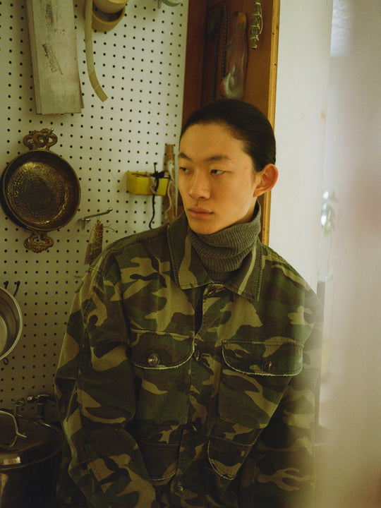 A person with long hair wearing a Camouflage Twill Utility Cargo Jacket by FOUND stands by a pegboard decorated with tools and a brass pan.