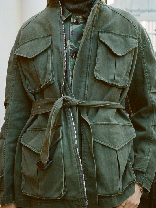 A person wears the FOUND M65 Utility Twill Pocket Jacket, a vintage-look, military-inspired green jacket featuring large front pockets and a tied waist belt.