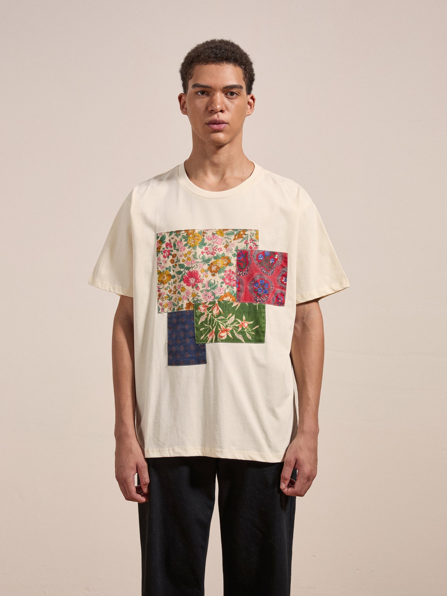 A person stands against a plain backdrop wearing the FOUND Patchwork Tee, which features a colorful patchwork design on the front, teamed with black pants and maintaining a neutral expression.