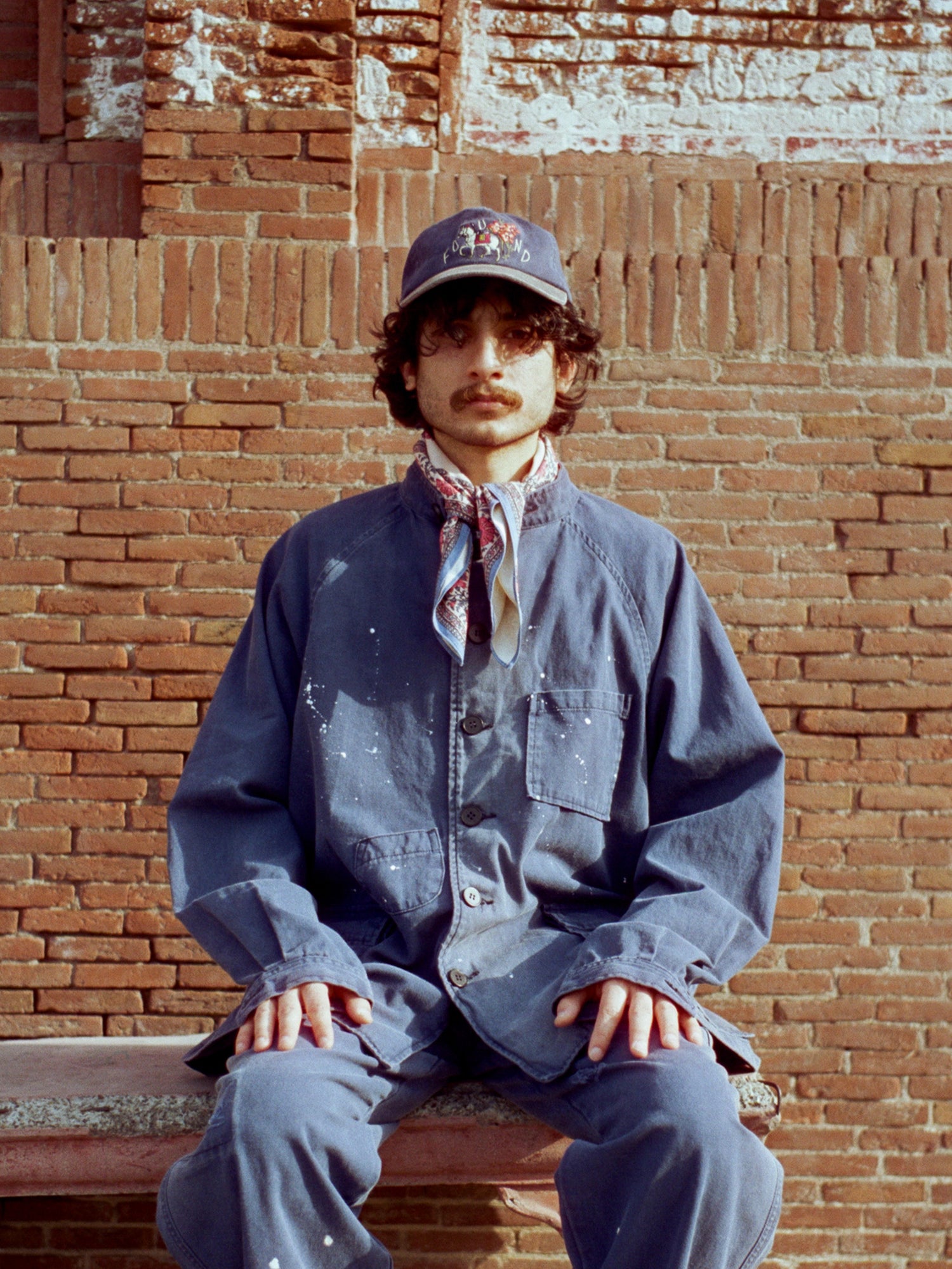 A man dressed in a River Painters Chore Coat by Found is sitting on a bench in front of a brick wall.