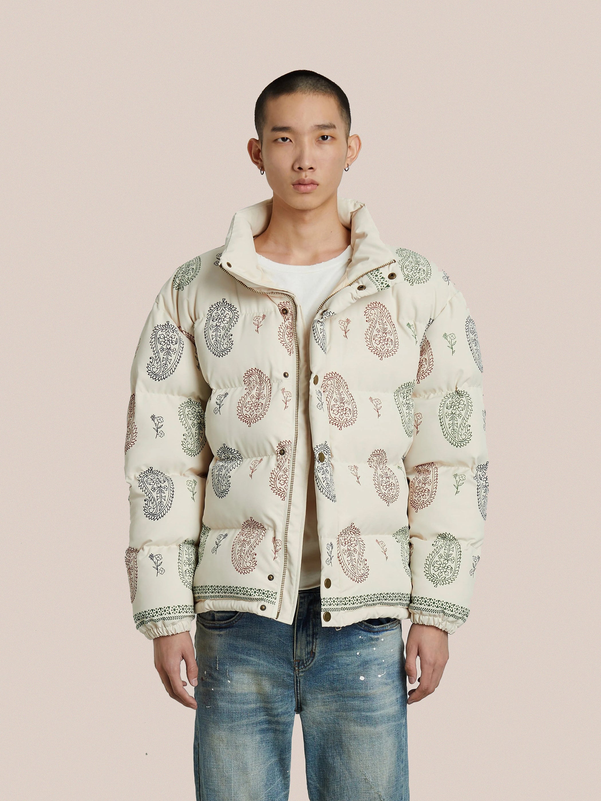 A person stands against a beige background wearing the FOUND Paisley Puffer Jacket in Cream with adjustable waist toggles, paired with jeans and an air of confidence as they look forward.